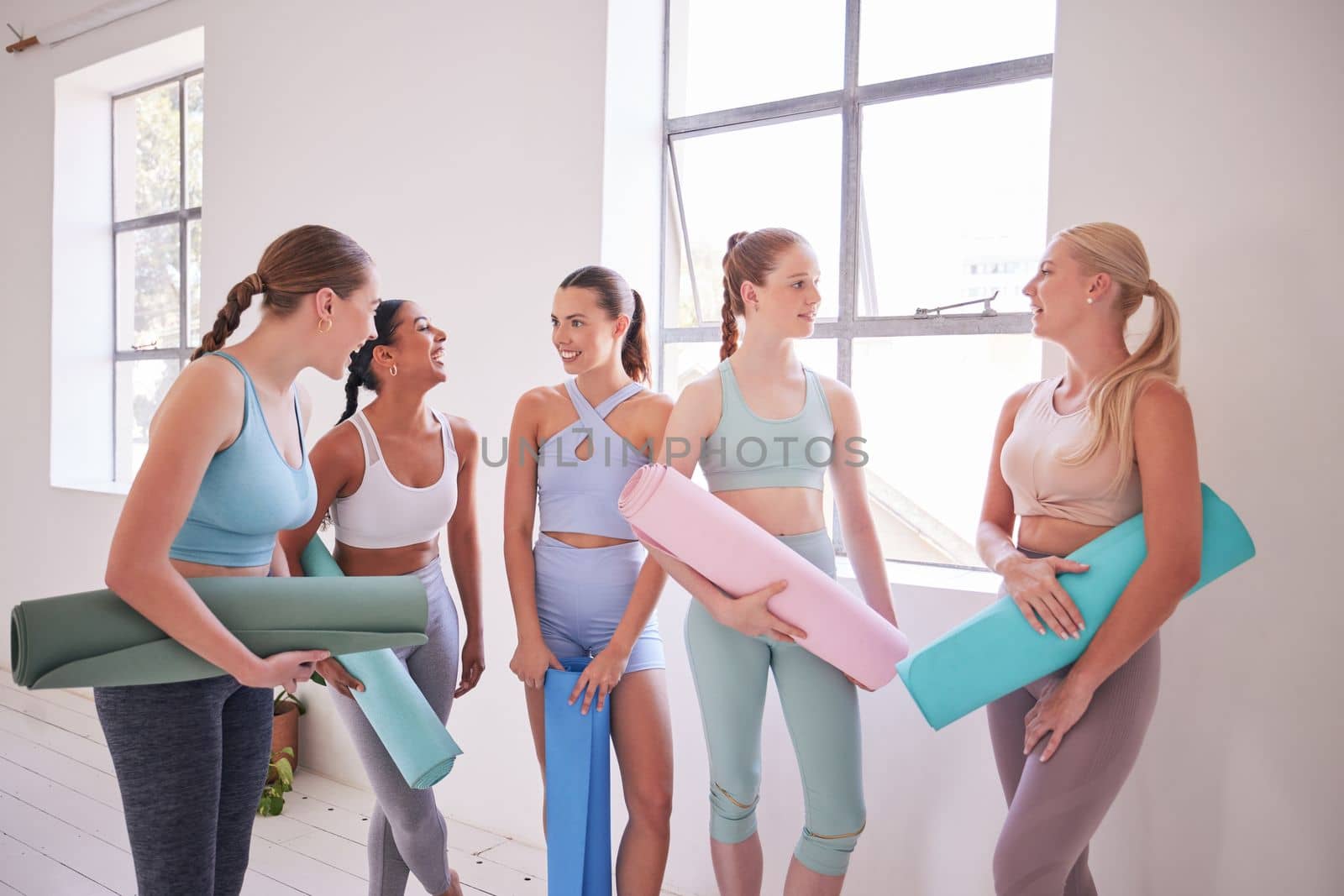 Friends talking before yoga class. Young women standing a pilates studio. Dedicated friends bonding before yoga class. Women waiting before holistic yoga class. Happy friends talking in yoga class by YuriArcurs