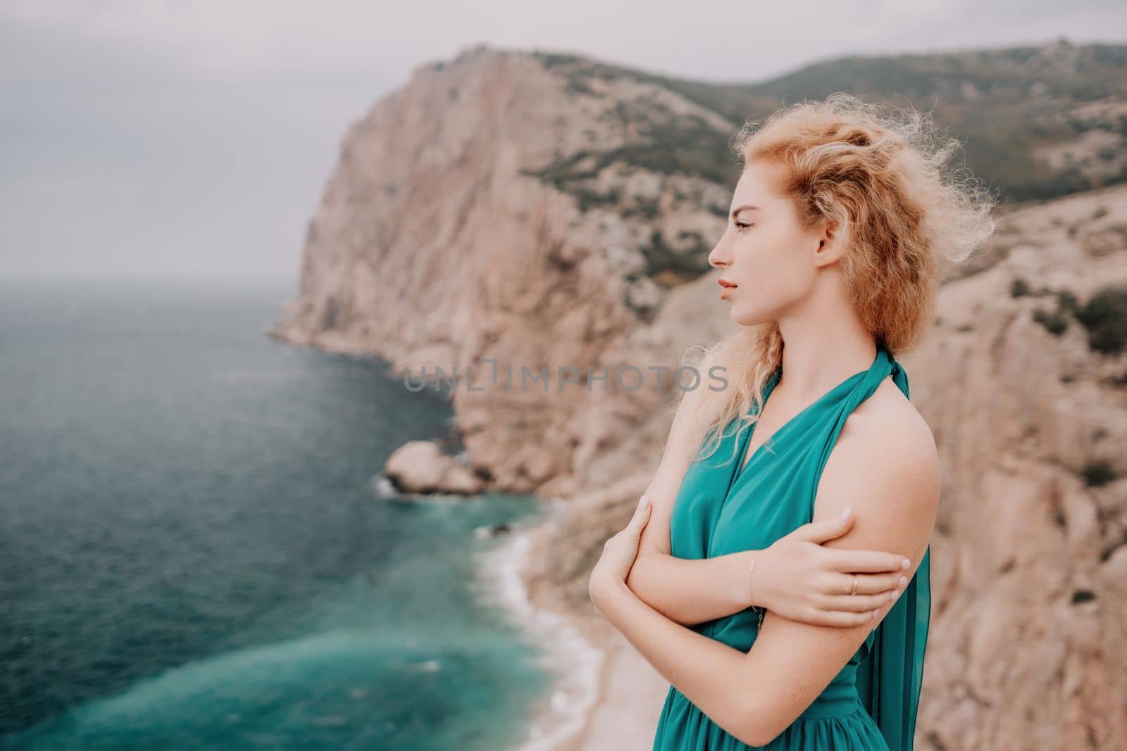 Side view a Young beautiful sensual woman in a mint long dress posing on a volcanic rock high above the sea during sunset. Girl on the nature on overcast sky background. Fashion photo