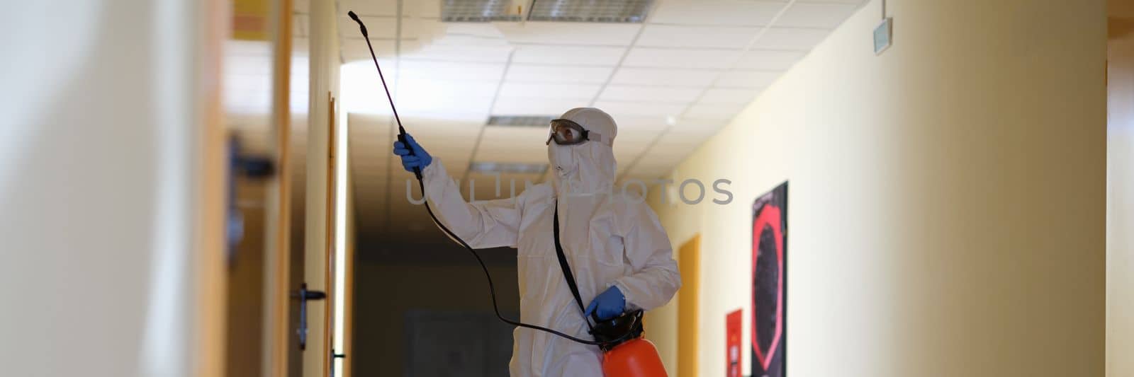 Person in protective suit with disinfectant cleaning public area in building during pandemic by kuprevich