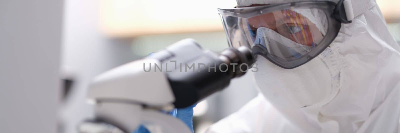 Biologist in protective suit with goggles and medical mask working with microscope in laboratory by kuprevich