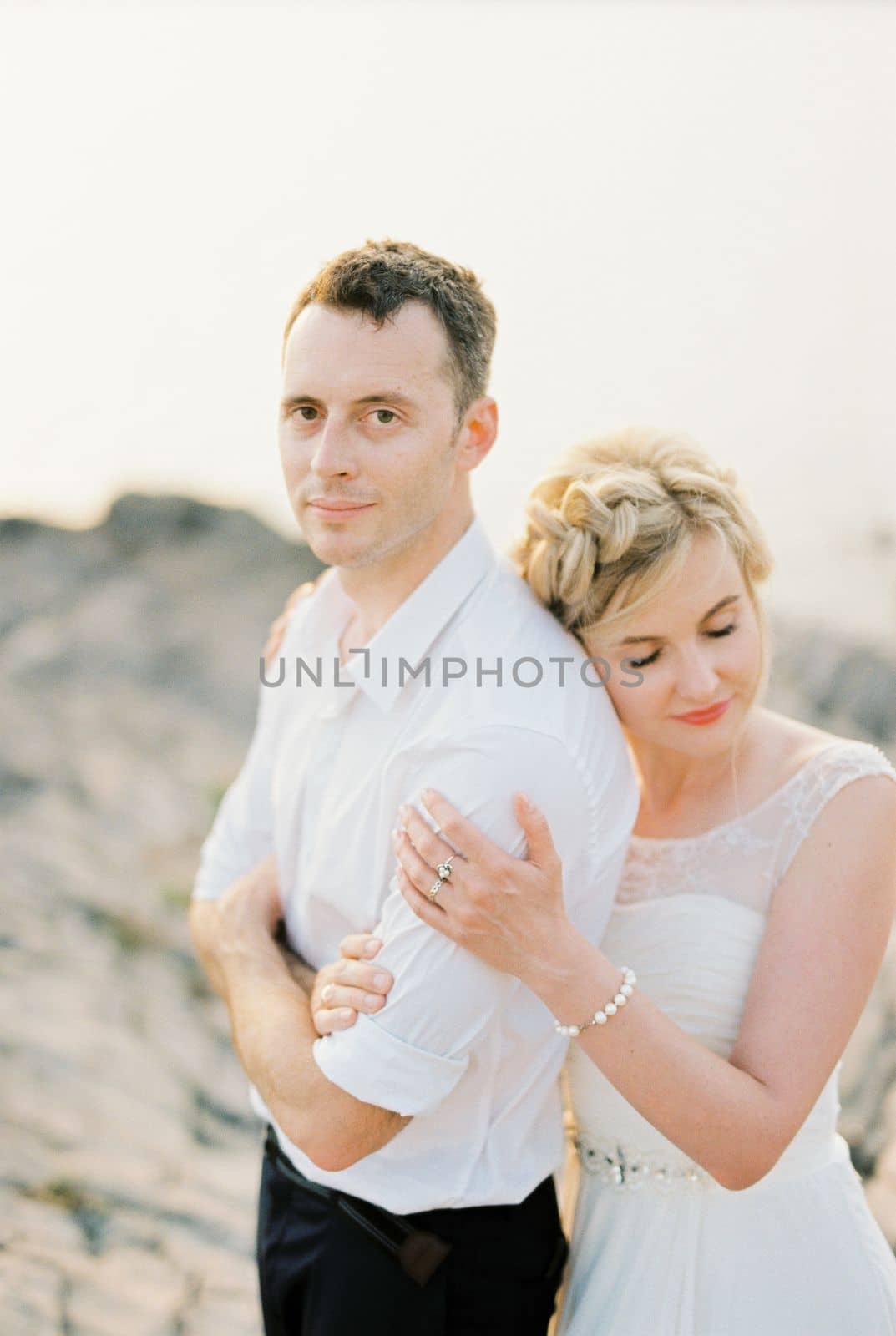 Bride hugs groom from behind with her head resting on his shoulder. Portrait by Nadtochiy