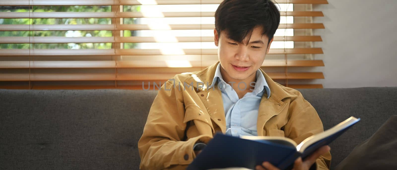 Pleased asian man sitting on comfortable sofa and reading interesting book, enjoying leisure weekend time at home by prathanchorruangsak