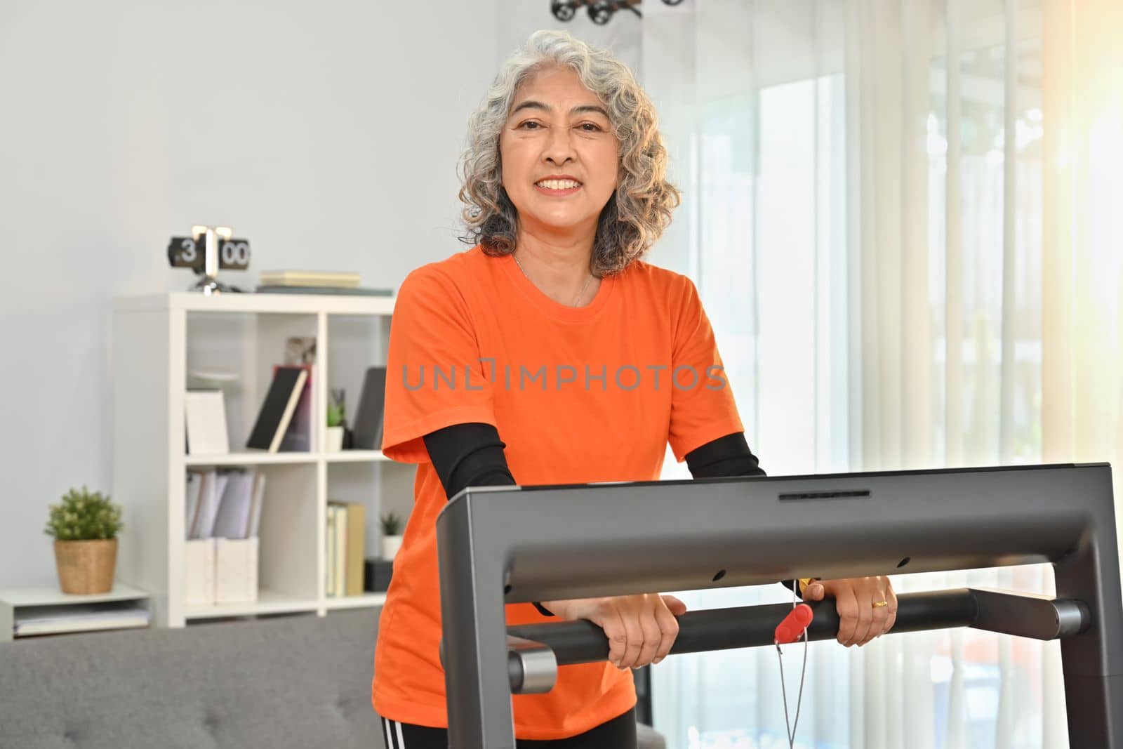 Smiling middle aged woman in sport clothes walking on a treadmill indoors. Healthy active lifestyle concept by prathanchorruangsak