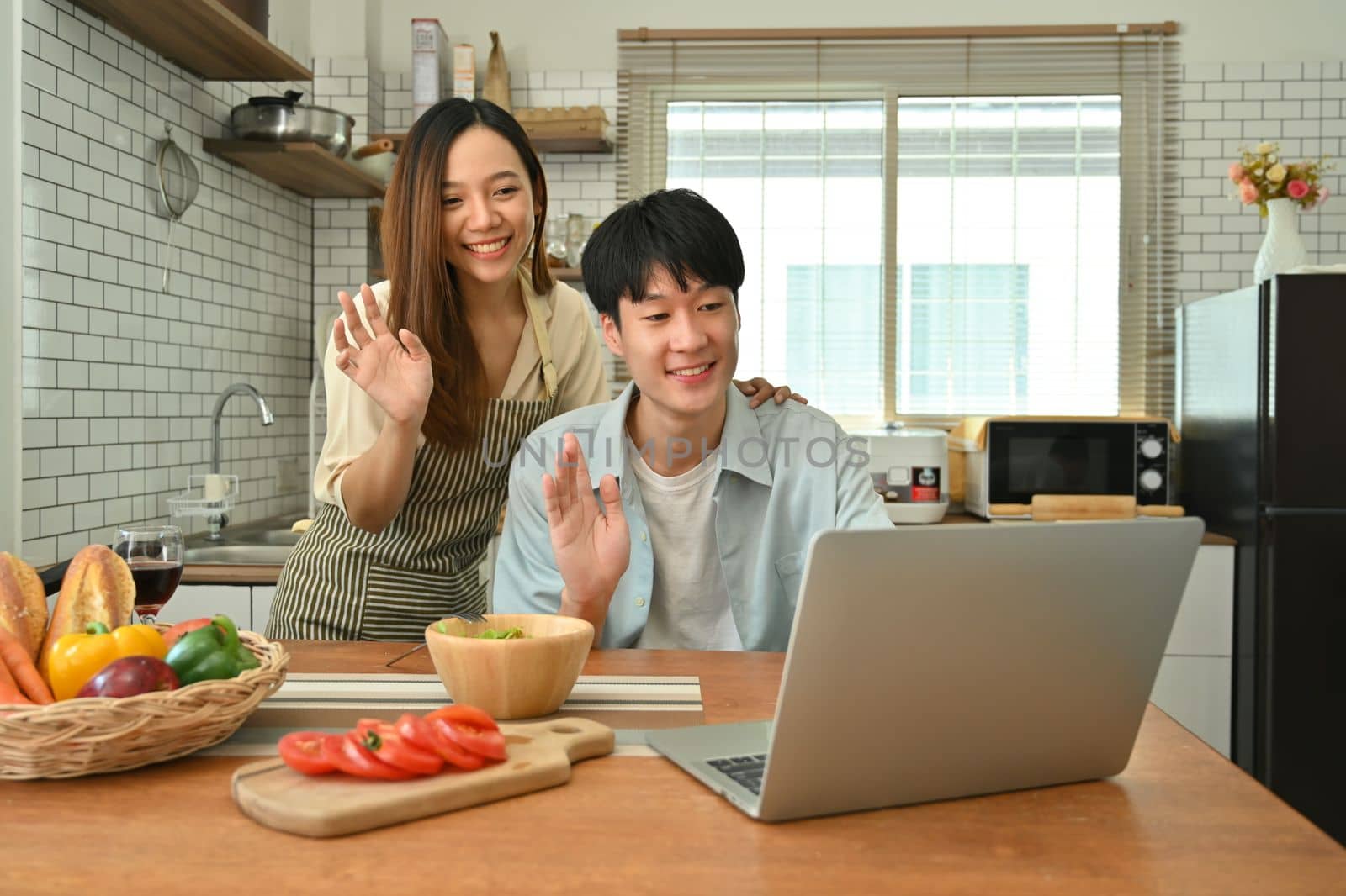 Smiling asian man and wife making video call, chatting online with family or friends on laptop in modern kitchen.
