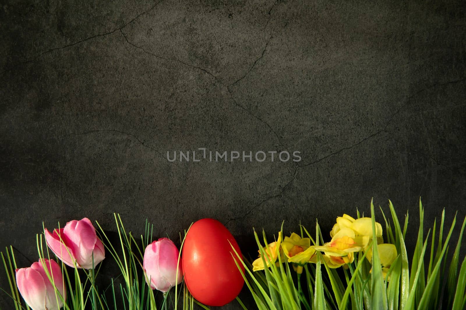 Easter eggs on fresh green grass against grey concrete background, colorful pink tulips and yellow daisies. Happy Easter holiday concept with copy space top view design egg hunting