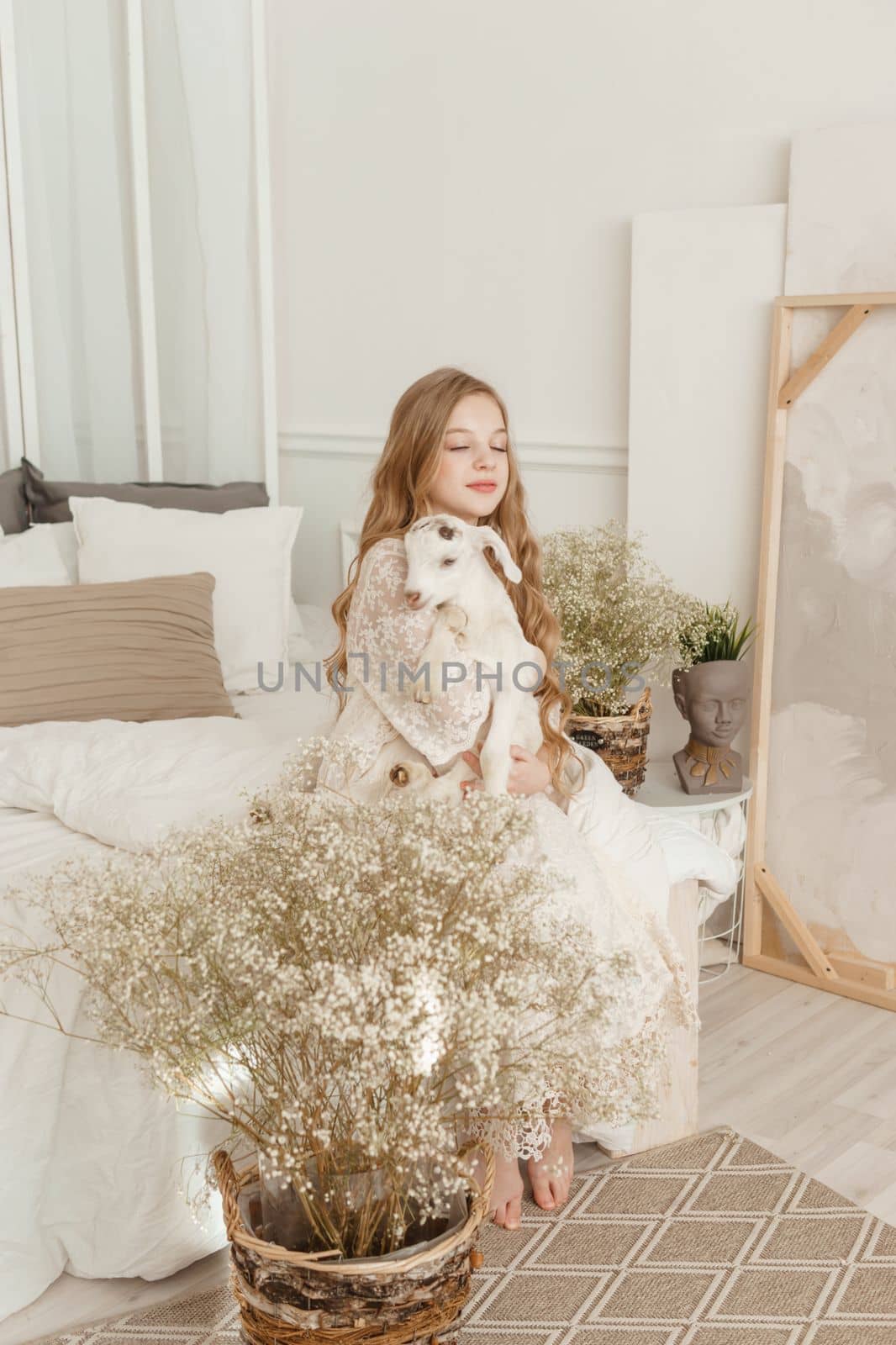 A blonde girl with long hair is sitting on the couch with a goat at home. The concept of animal care