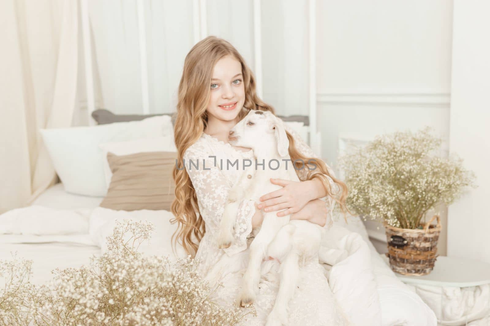 A blonde girl with long hair is sitting on the couch with a goat at home. The concept of animal care