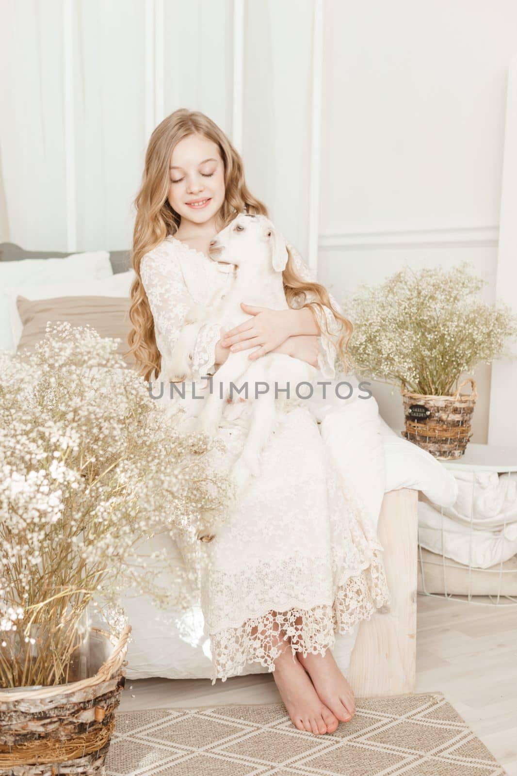 A blonde girl with long hair is sitting on the couch with a goat at home. The concept of animal care
