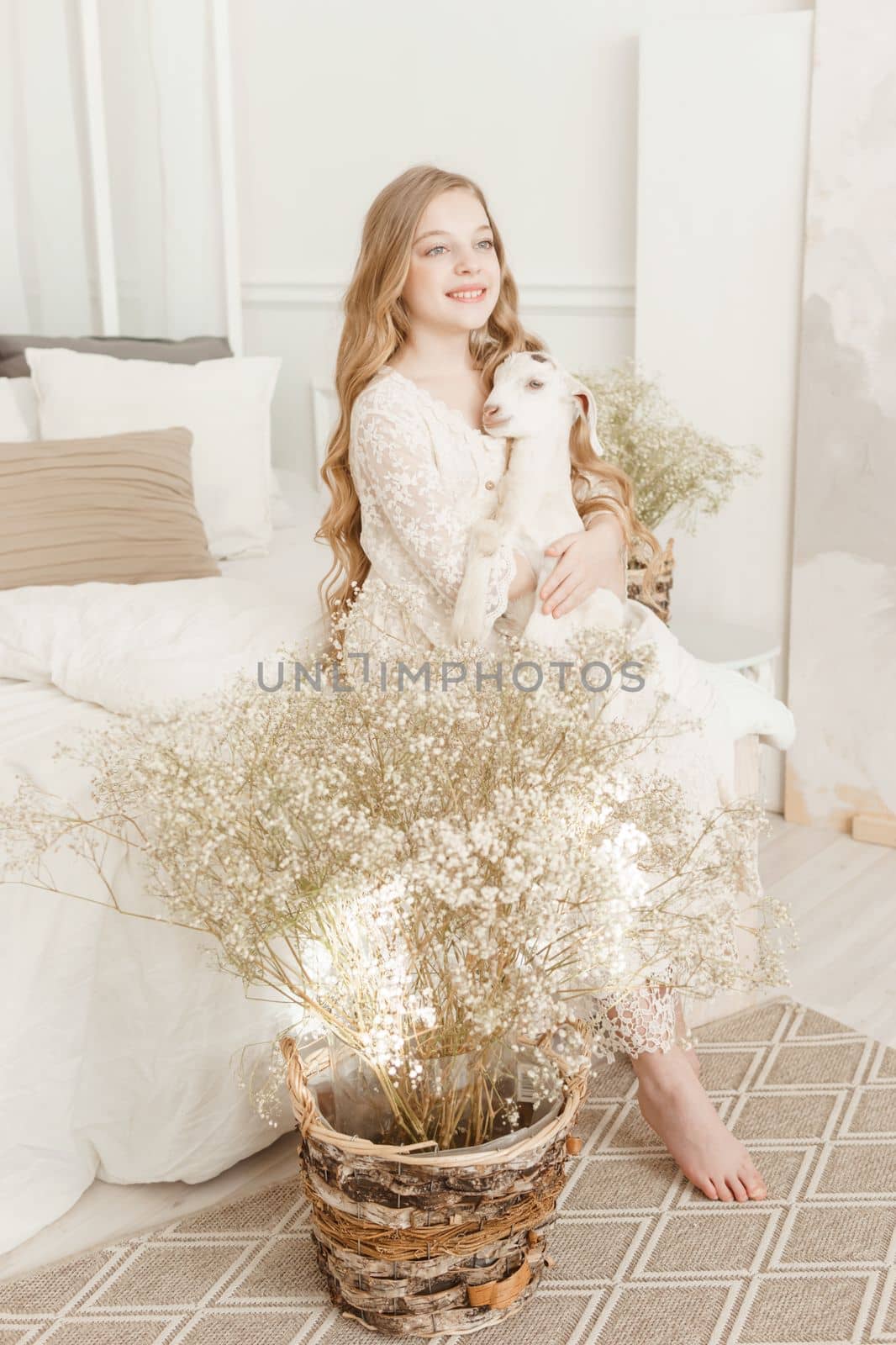 A blonde girl with long hair is sitting on the couch with a goat at home. The concept of animal care