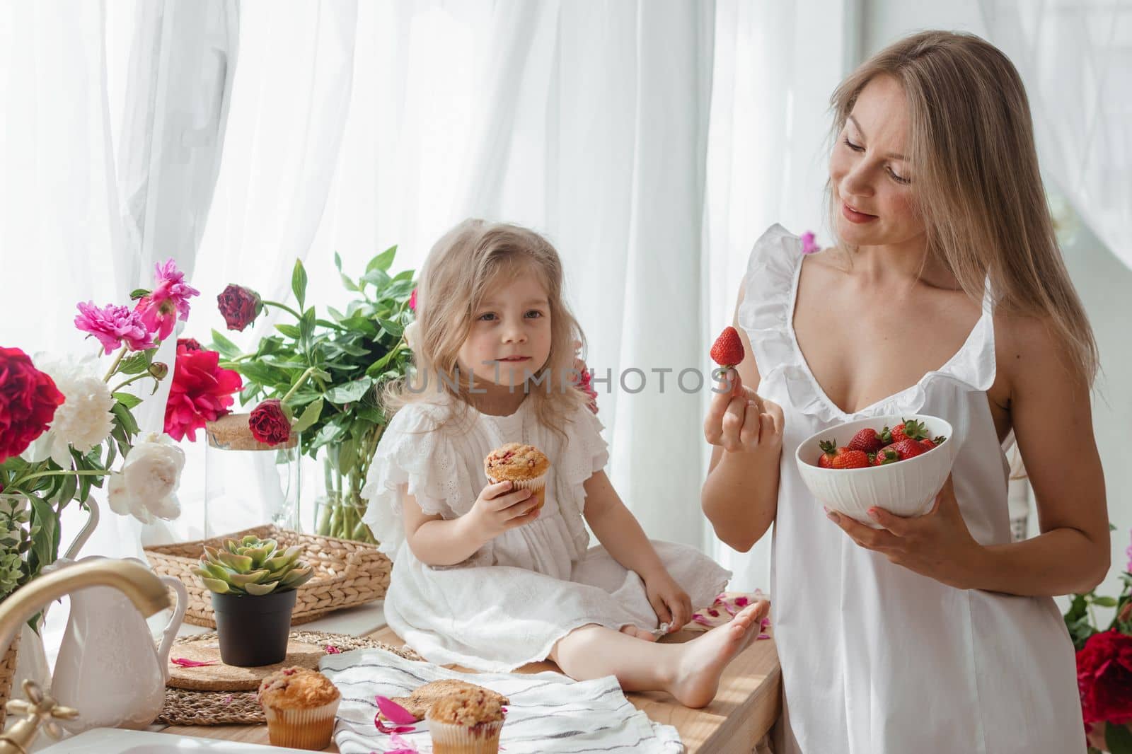 A little blonde girl with her mom on a kitchen countertop decorated with peonies. The concept of the relationship between mother and daughter. Spring atmosphere.