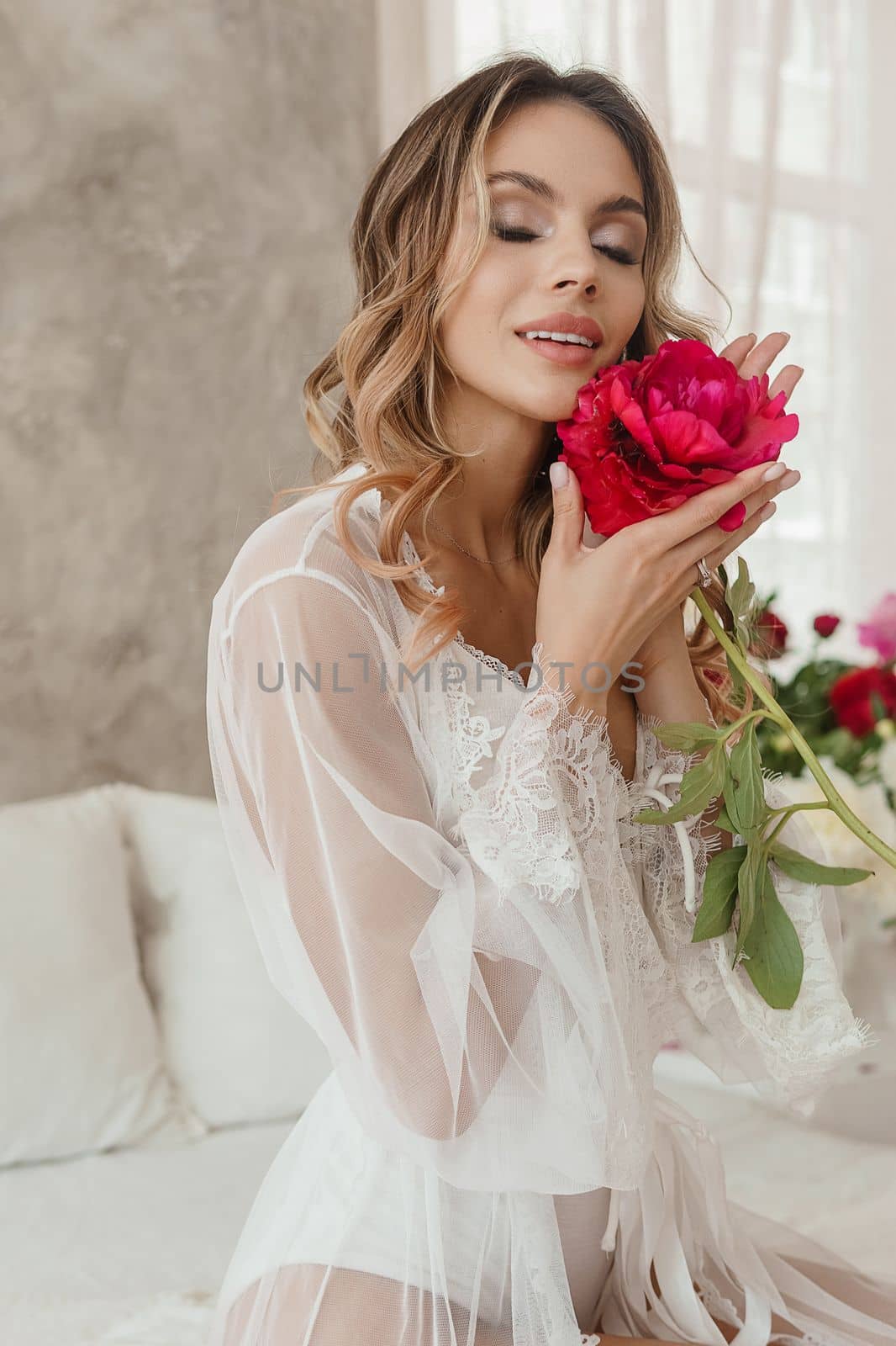 A beautiful girl in a white nightgown at home, surrounded by spring flowers on a large bed. A room decorated with bouquets of peonies.