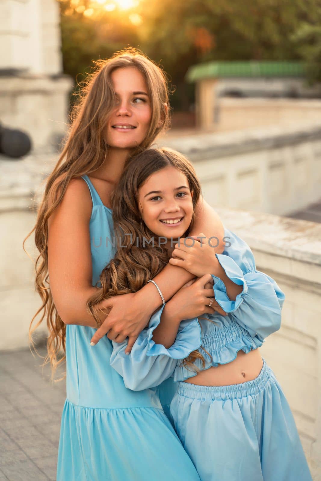 Portrait of mother and daughter in blue dresses with flowing long hair against the backdrop of sunset. The woman hugs and presses the girl to her. They are looking at the camera