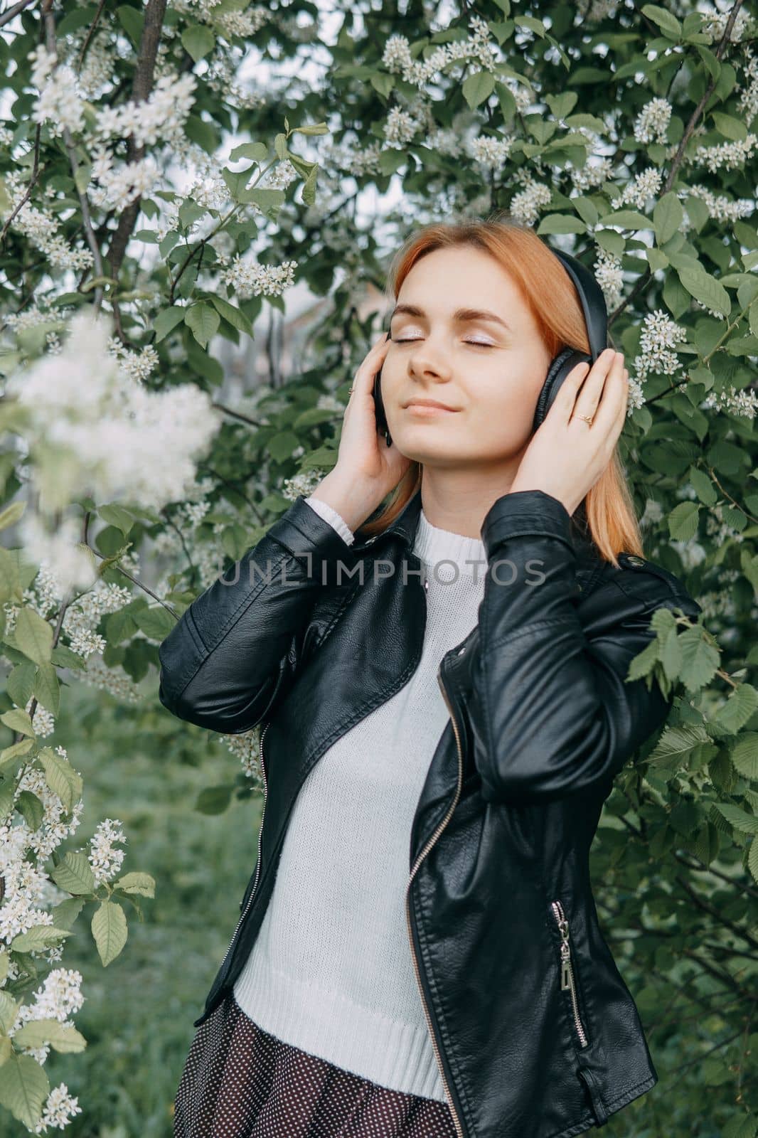A red-haired teenage girl listens to music in headphones, configured via the phone. The time of spring flowering of cherry orchards.