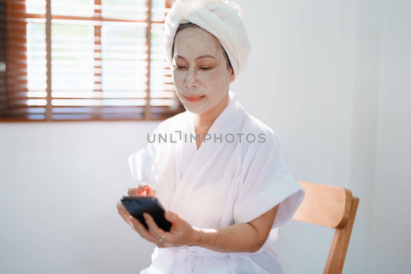 Portrait of elderly woman doing face spa treatment and using mobile phone. by Manastrong