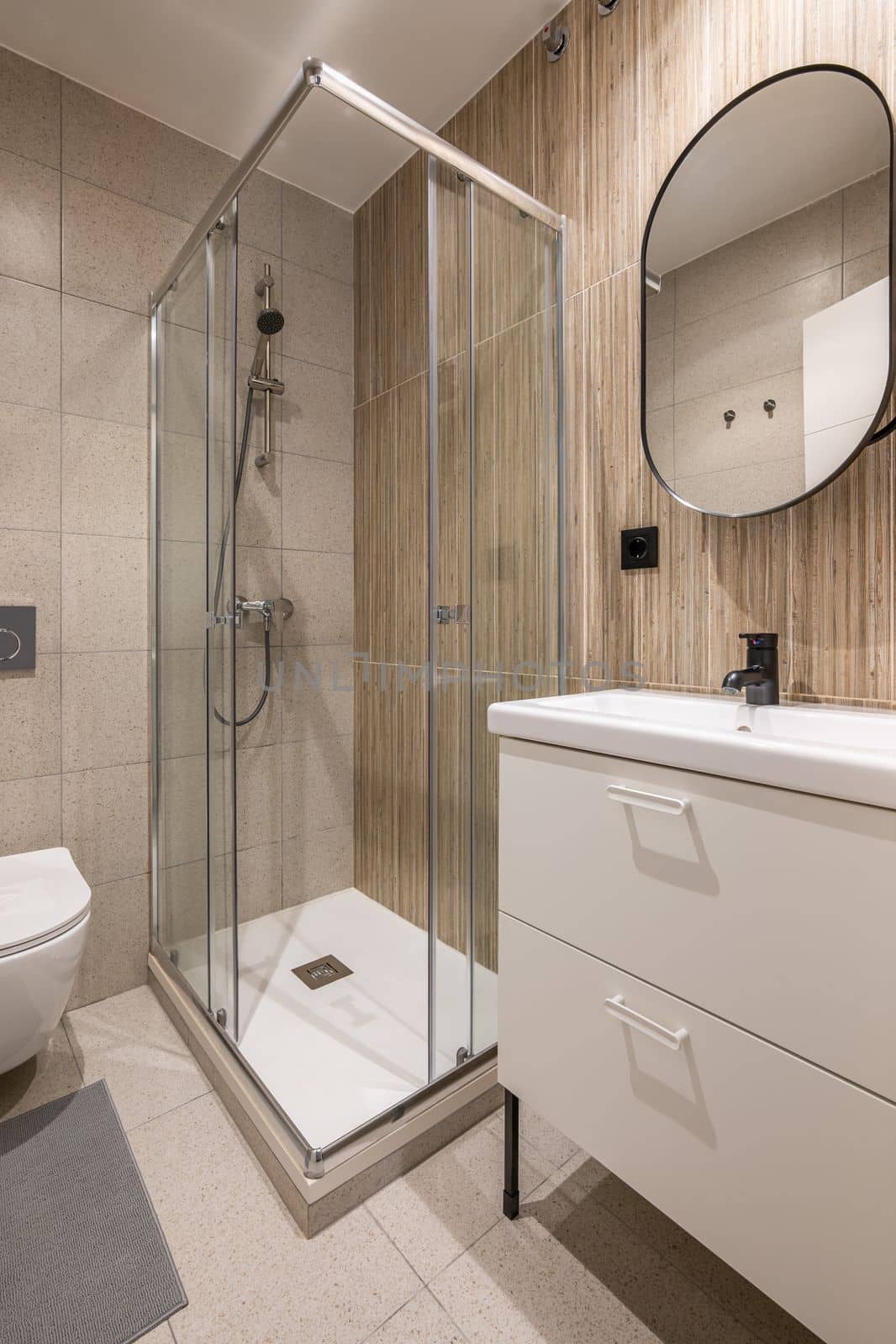 Bathroom with large shower area in corner. Rectangular washbasin on vanity table with white drawers and mirror on wall in black metal frame. Walls made of granite tiles of different textures