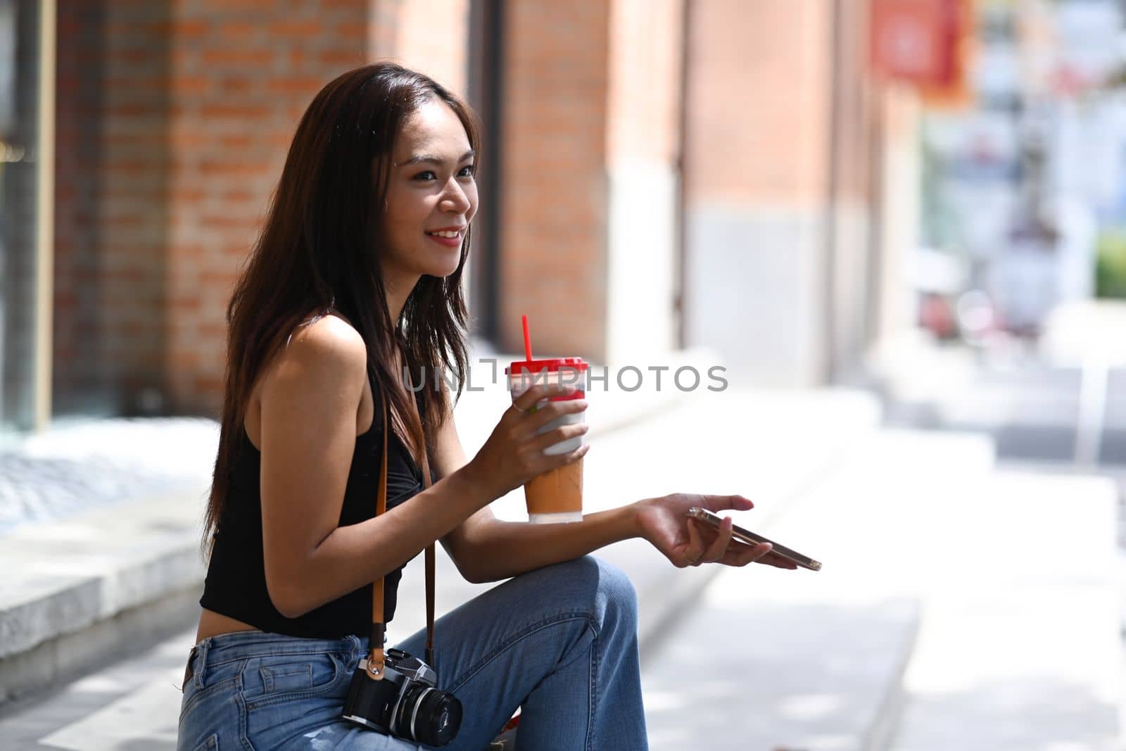 Smiling young woman sitting outdoor holding ice coffee and using smart phone.