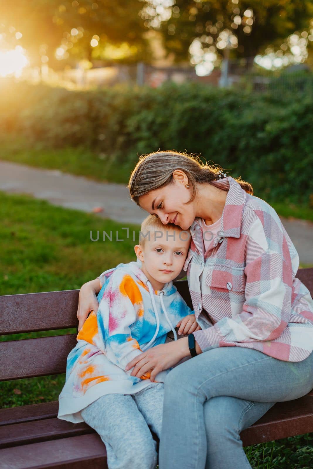 mother and son sit on a park bench in the rays of the setting sun. the concept of a family. Mother's Day. beautiful girl (mother) with a boy (son) in the park in the park are sitting on a bench at sunset.