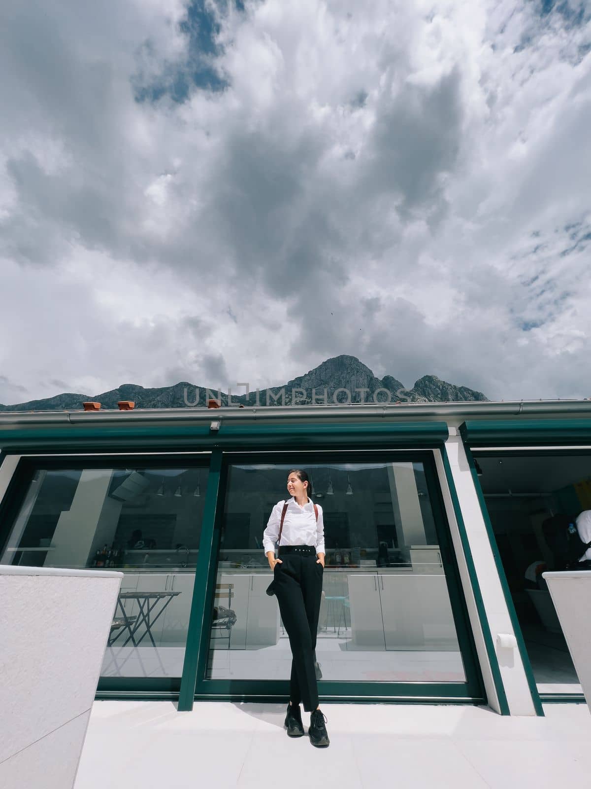 Young woman in a white shirt and black trousers stands near the panoramic window of the hotel. High quality photo