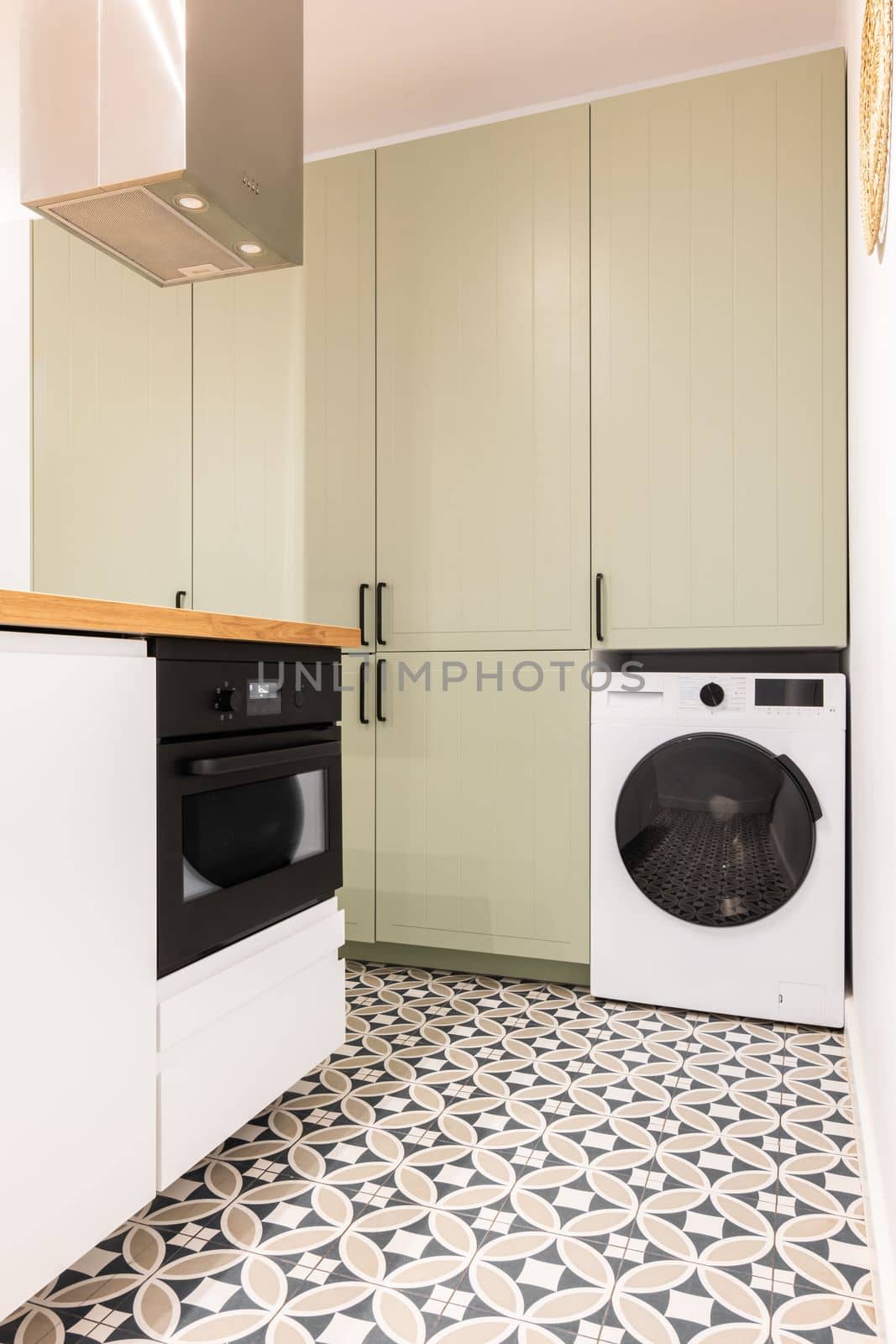 Part of spacious kitchen with modern appliances with black elements. Large built-in wooden cabinet for kitchenware and utensils in beige color with black handles. Floor is made of marble mosaic tiles. by apavlin