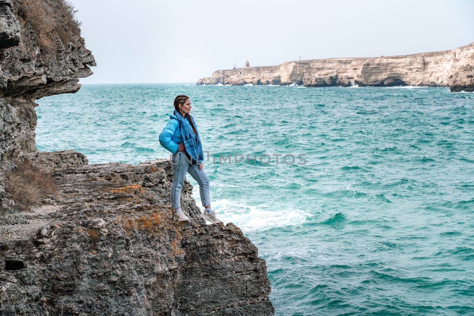 A woman in a blue jacket stands on a rock above a cliff above the sea and looks at the raging ocean. Girl traveler rests, thinks, dreams, enjoys nature. Peace and calm landscape, windy weather