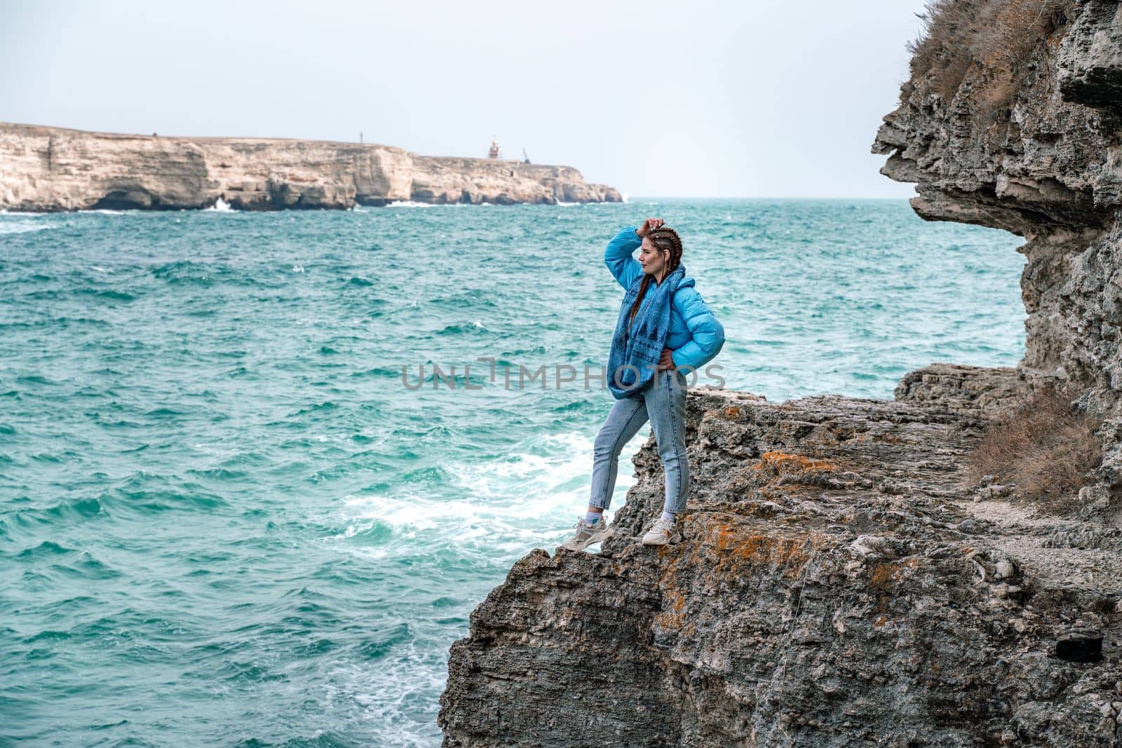 A woman in a blue jacket stands on a rock above a cliff above the sea and looks at the raging ocean. Girl traveler rests, thinks, dreams, enjoys nature. Peace and calm landscape, windy weather. by Matiunina