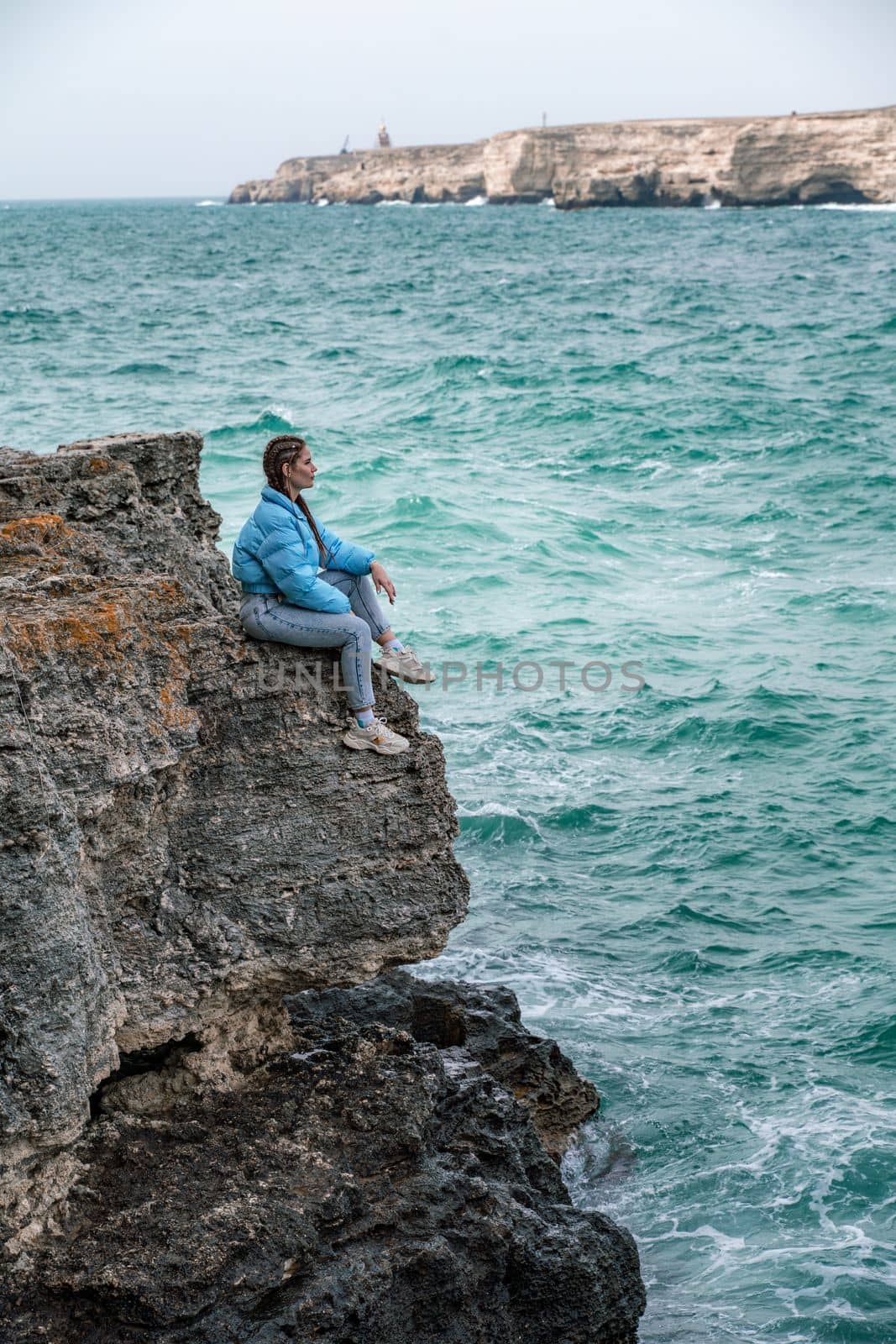 Woman rocks sea. A woman in a blue jacket sits on a rock above a rock above the sea and looks at the raging ocean. Girl traveler rests, thinks, dreams, enjoys nature