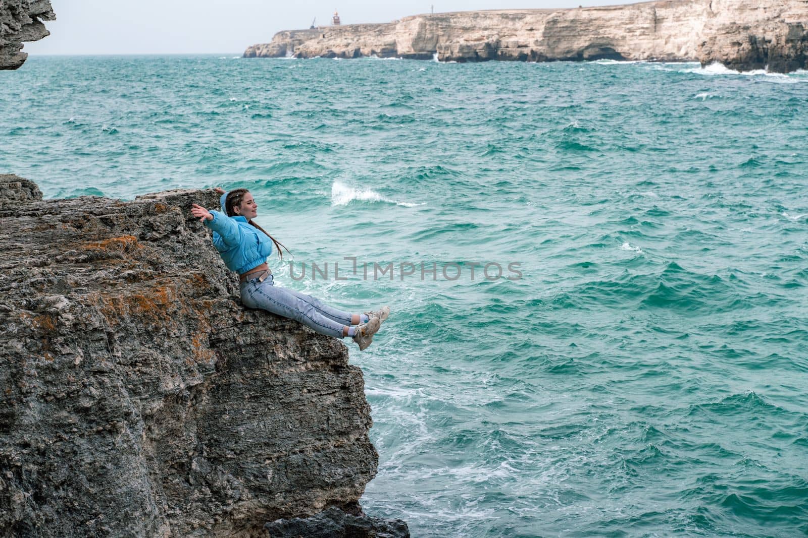 A woman in a blue jacket sits on a rock above a cliff above the sea, looking at the stormy ocean. Girl traveler rests, thinks, dreams, enjoys nature. Peace and calm landscape, windy weather