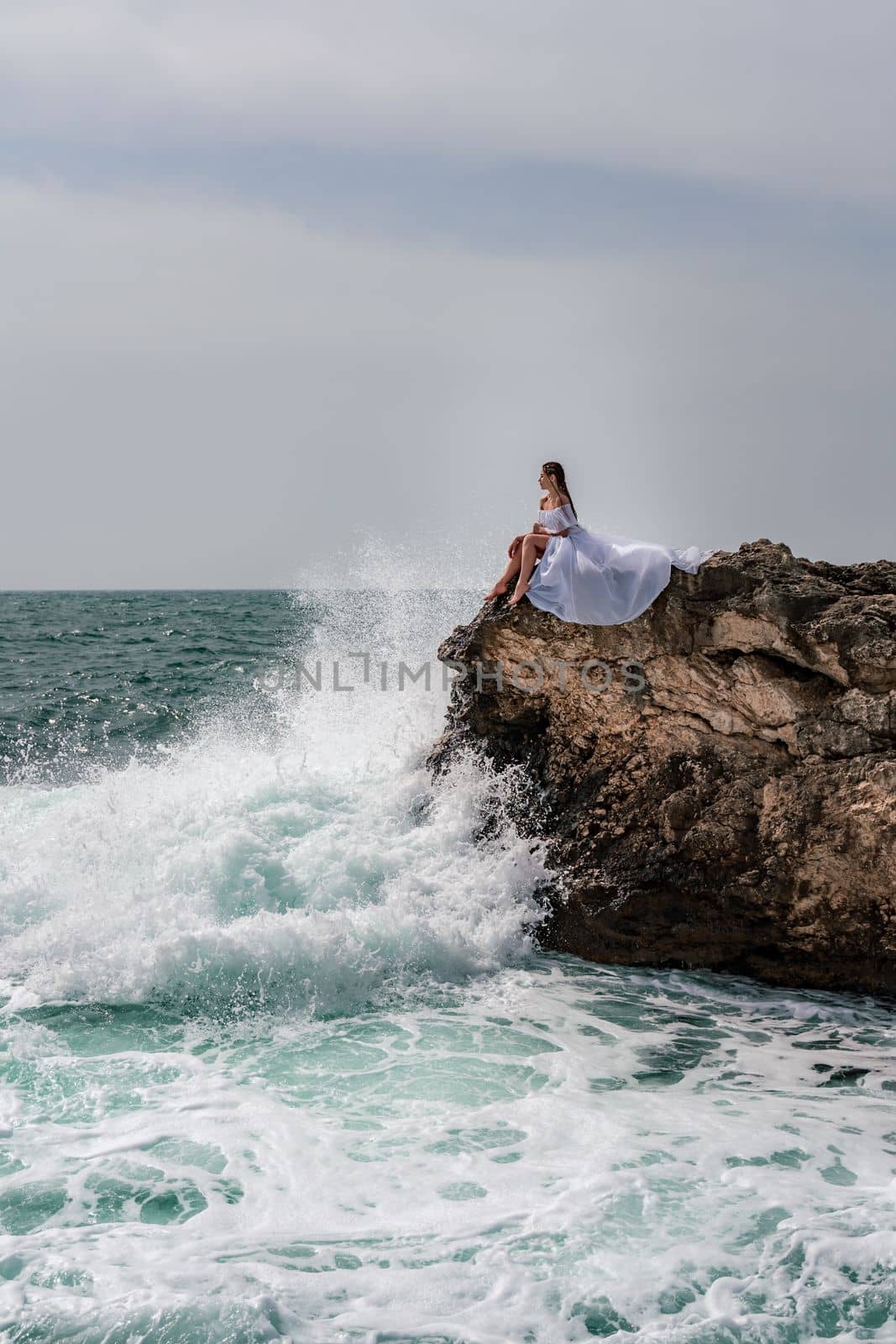 A woman in a storm sits on a stone in the sea. Dressed in a white long dress, waves crash against the rocks and white spray rises above her