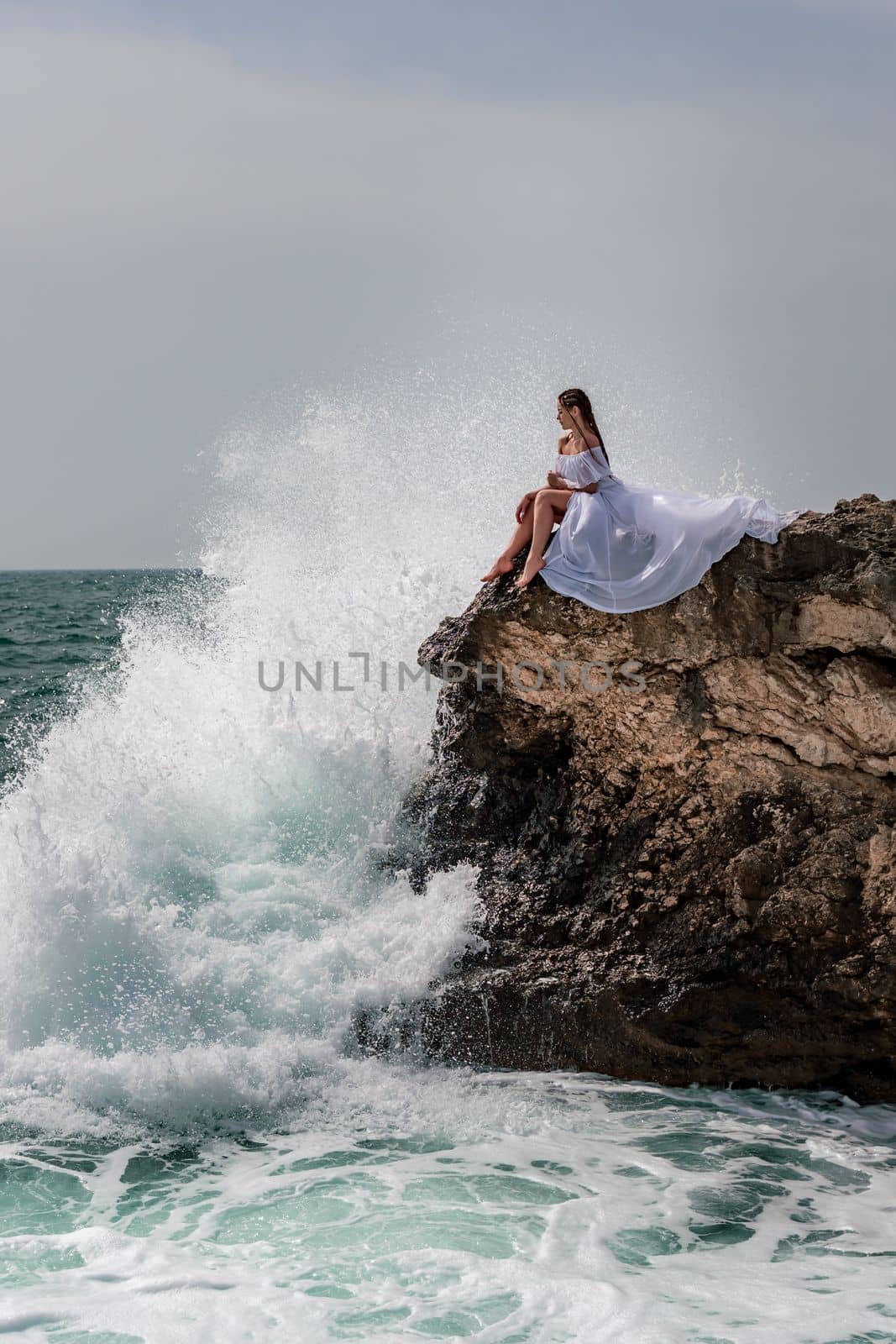 A woman in a storm sits on a stone in the sea. Dressed in a white long dress, waves crash against the rocks and white spray rises above her. by Matiunina