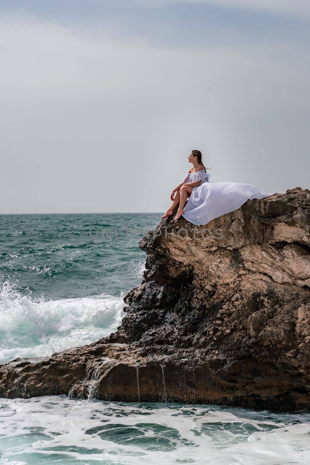 A woman in a storm sits on a stone in the sea. Dressed in a white long dress, waves crash against the rocks and white spray rises. by Matiunina