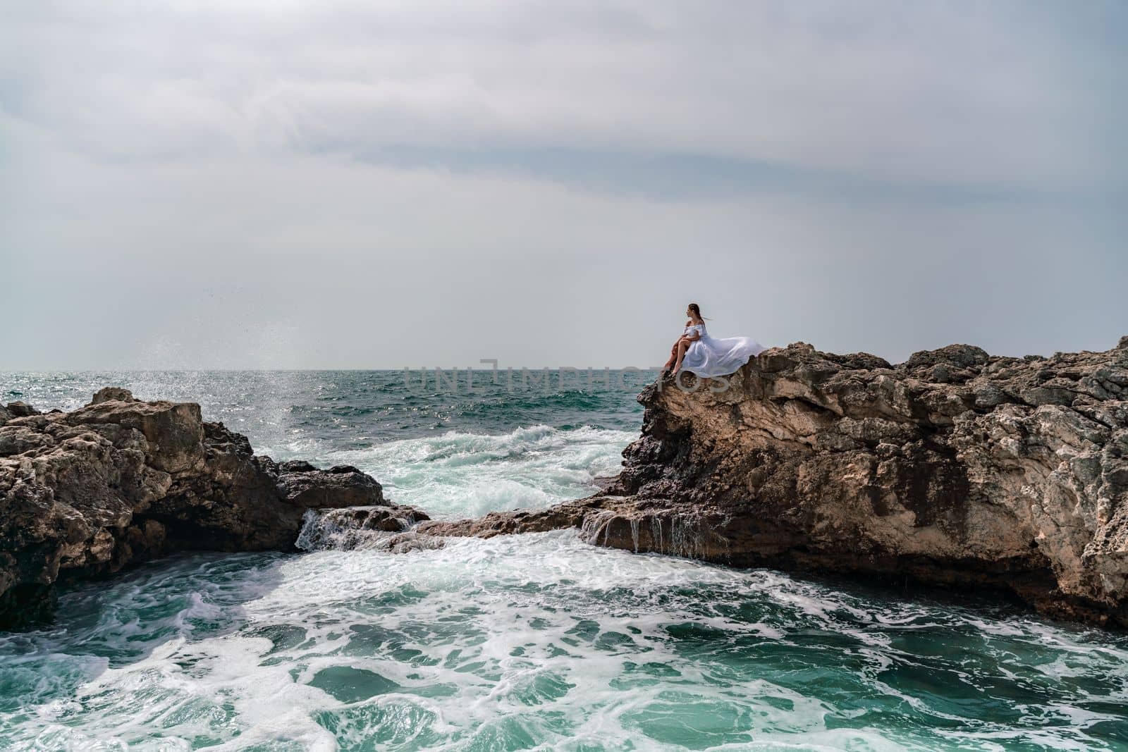 A woman in a storm sits on a stone in the sea. Dressed in a white long dress, waves crash against the rocks and white spray rises