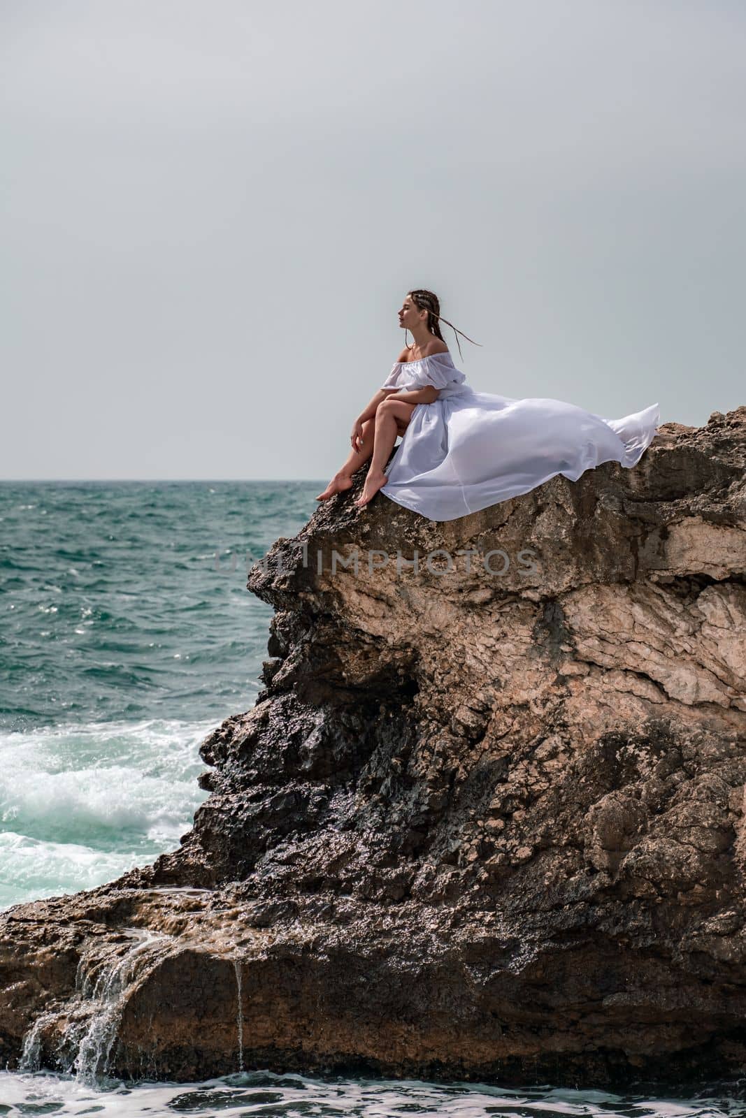 A woman in a storm sits on a stone in the sea. Dressed in a white long dress, waves crash against the rocks and white spray rises