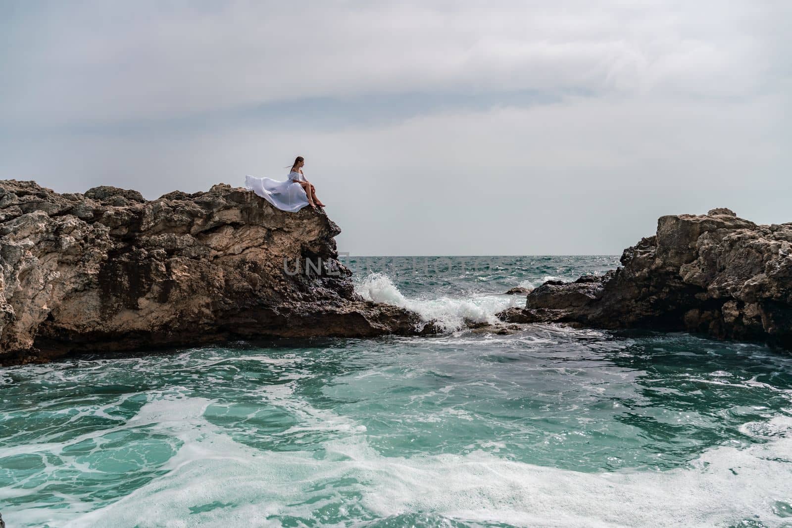 A woman in a storm sits on a stone in the sea. Dressed in a white long dress, waves crash against the rocks and white spray rises. by Matiunina