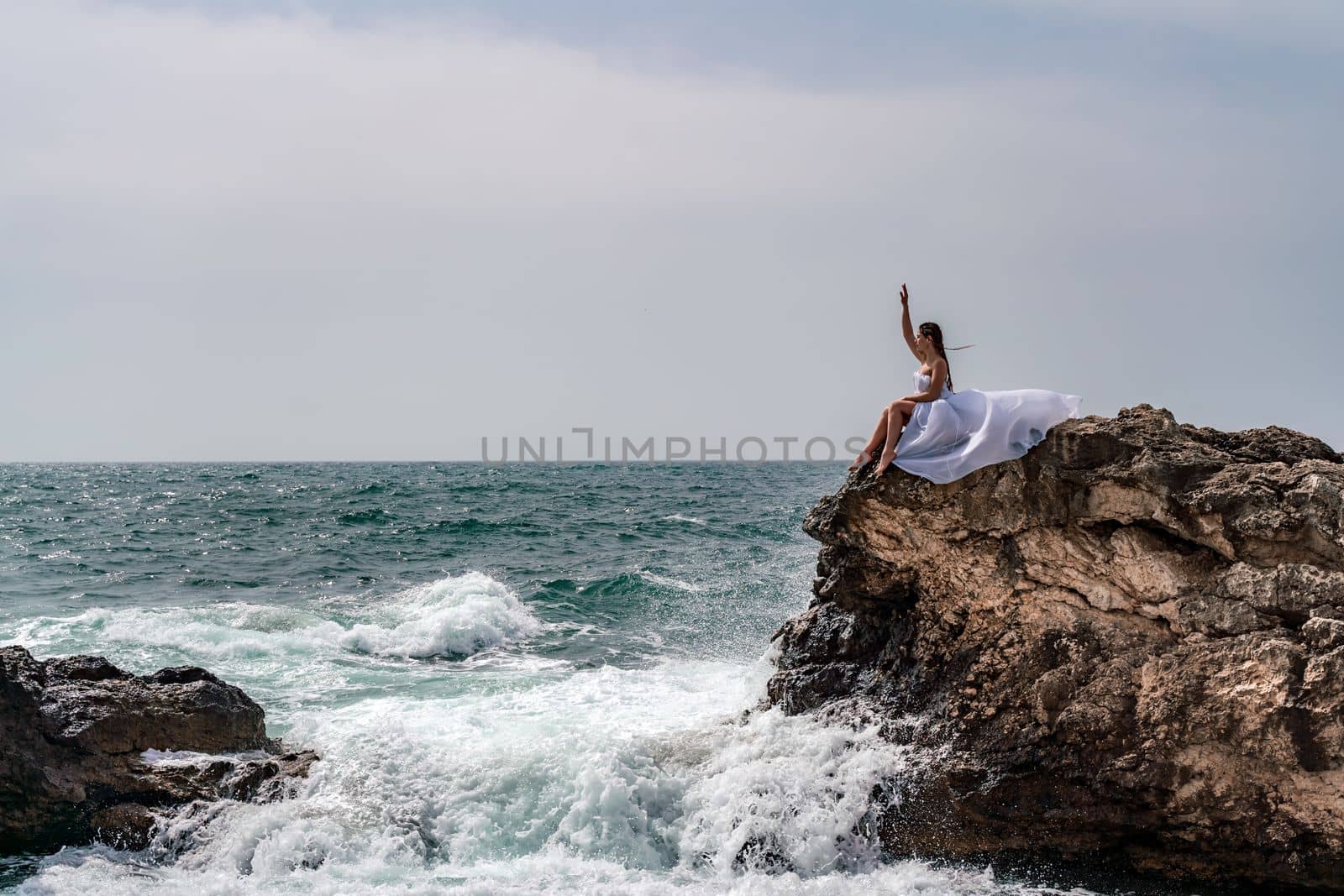A woman in a storm sits on a stone in the sea. Dressed in a white long dress, waves crash against the rocks and white spray rises. by Matiunina