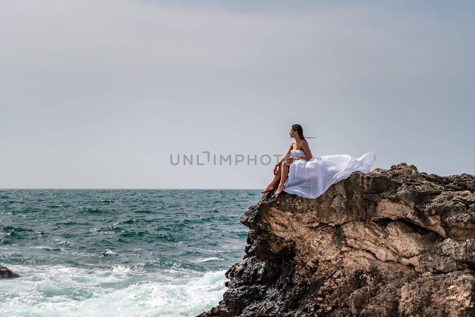 A woman in a storm sits on a stone in the sea. Dressed in a white long dress, waves crash against the rocks and white spray rises