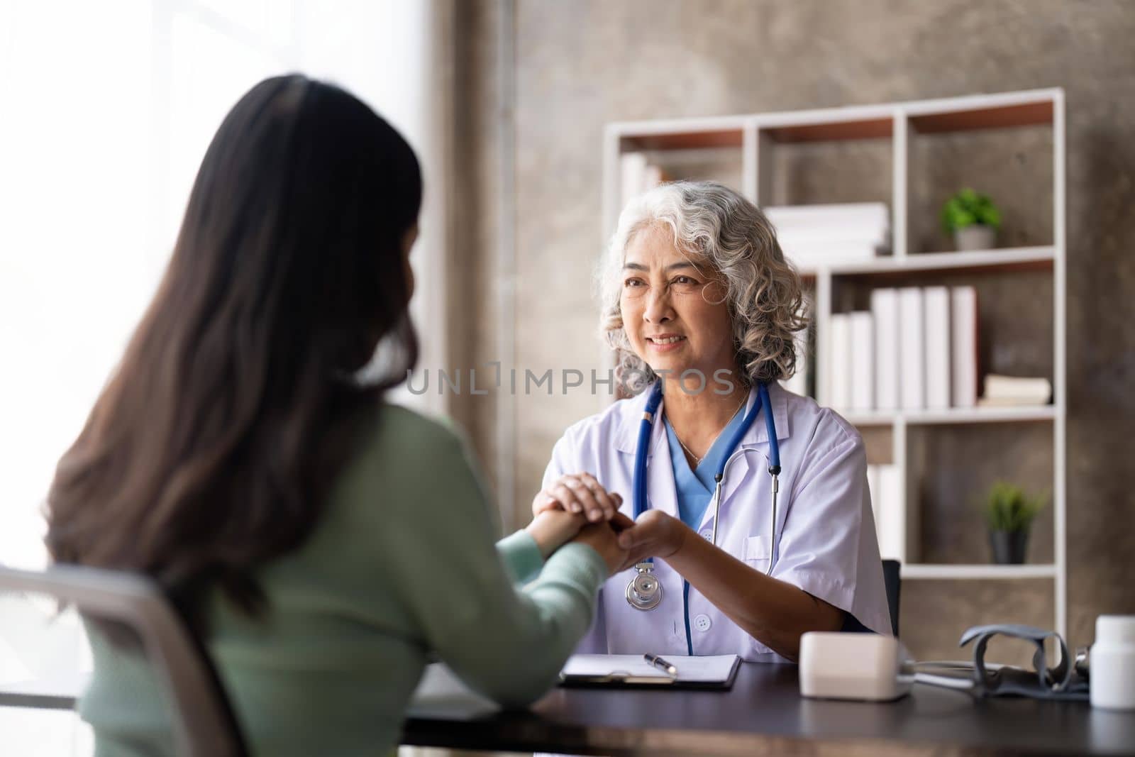 Woman senior doctor is Reading Medical History of Female Patient and Speaking with Her During Consultation in a Health Clinic. Physician in Lab Coat Sitting Behind a Laptop in Hospital Office by nateemee