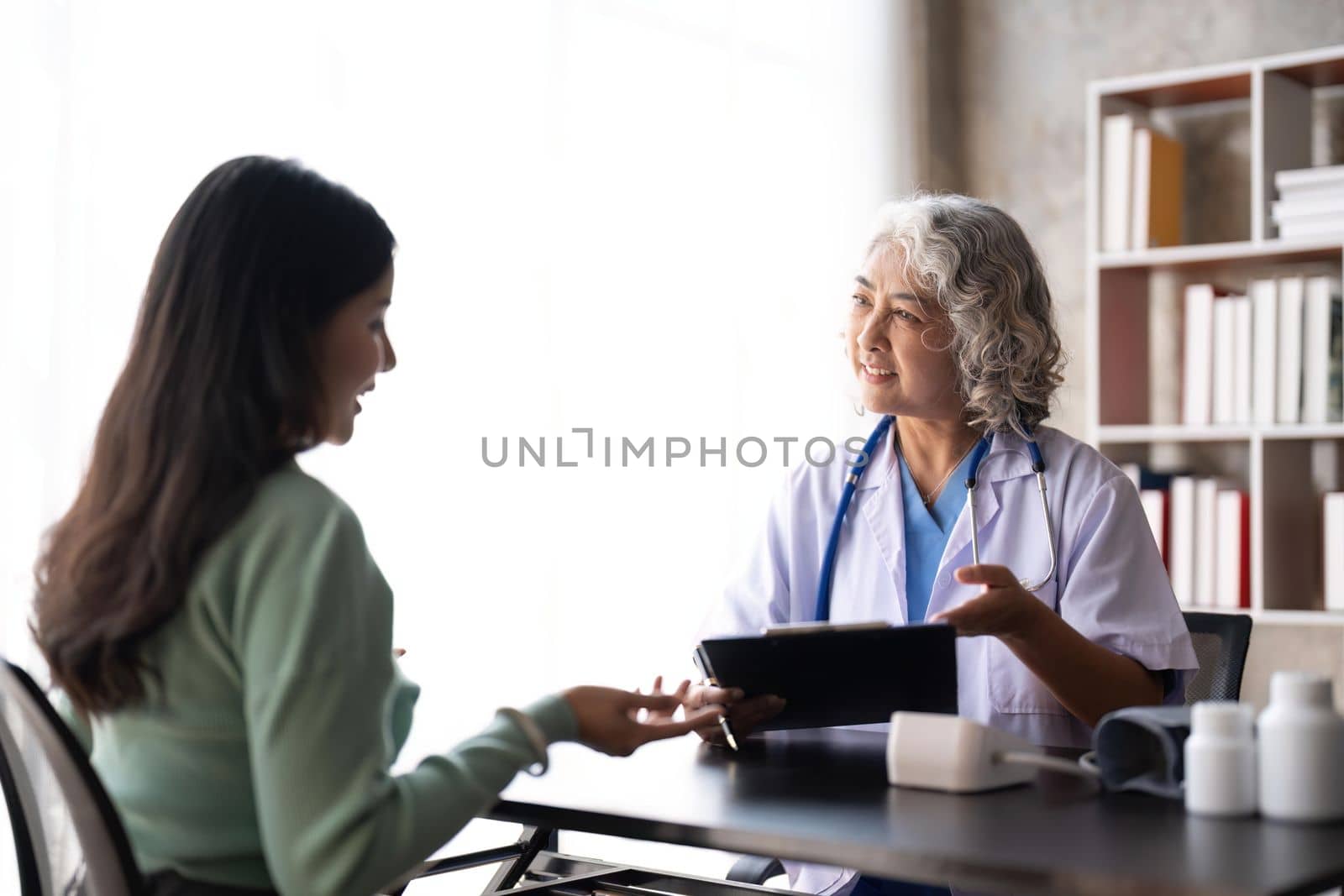 Woman senior doctor is Reading Medical History of Female Patient and Speaking with Her During Consultation in a Health Clinic. Physician in Lab Coat Sitting Behind a Laptop in Hospital Office.