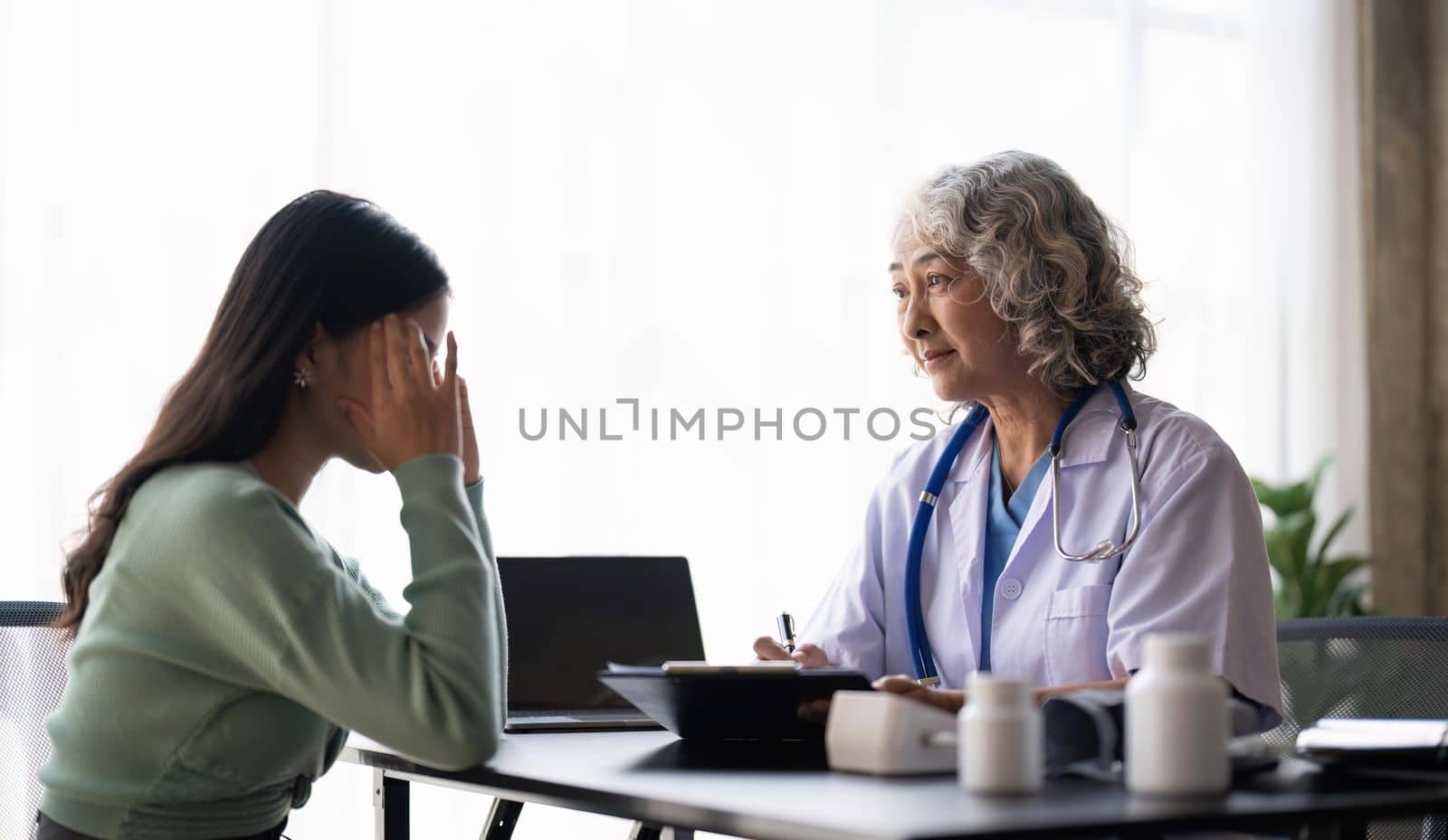 Woman senior doctor is Reading Medical History of Female Patient and Speaking with Her During Consultation in a Health Clinic. Physician in Lab Coat Sitting Behind a Laptop in Hospital Office by nateemee