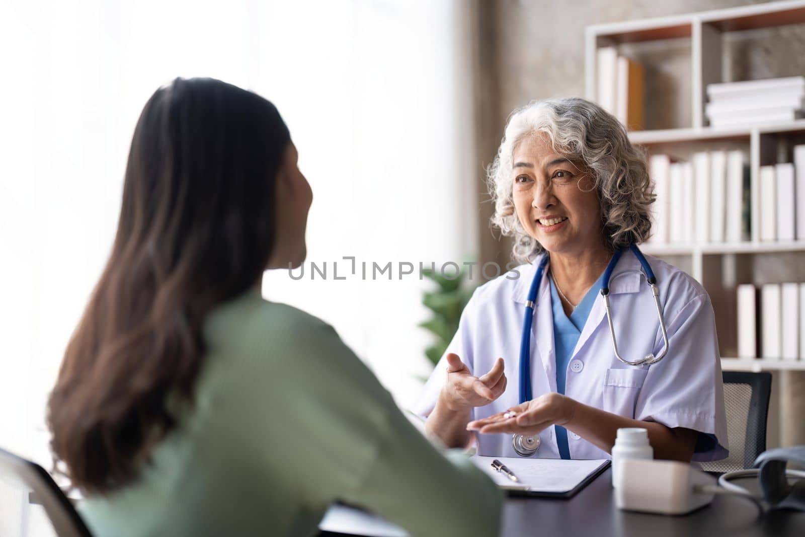 Woman senior doctor is Reading Medical History of Female Patient and Speaking with Her During Consultation in a Health Clinic. Physician in Lab Coat Sitting Behind a Laptop in Hospital Office by nateemee