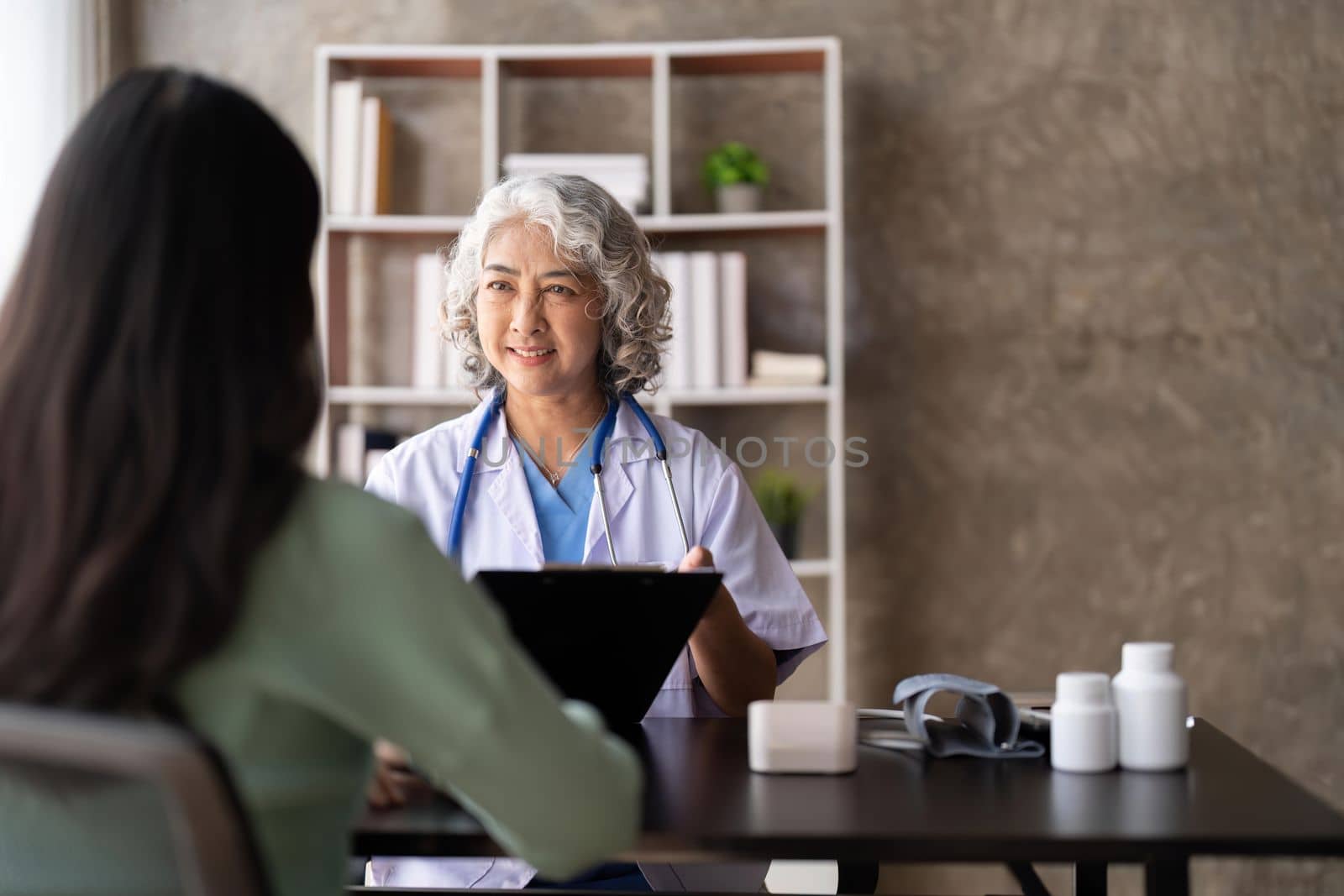 Woman senior doctor is Reading Medical History of Female Patient and Speaking with Her During Consultation in a Health Clinic. Physician in Lab Coat Sitting Behind a Laptop in Hospital Office.