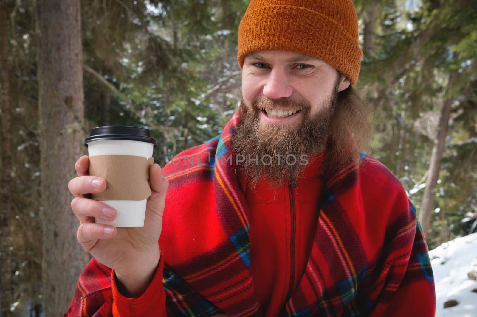Walk with a cup of hot cocoa in the winter park. Portrait of a handsome bearded guy in winter clothes in the snow drinking hot tea or coffee from a paper cup and enjoying a nice sunny snowy day by yanik88