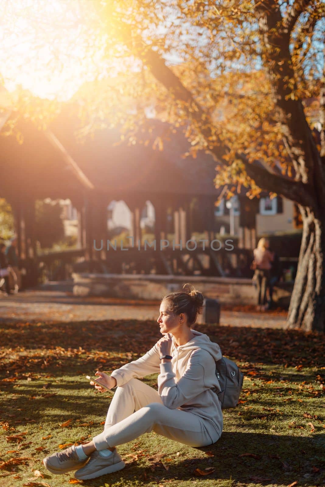 Young fashionable teenage girl with smartphone in park in autumn sitting at smiling. Trendy young woman in fall in park texting. Retouched, vibrant colors. Beautiful blonde teenage girl wearing casual modern autumn outfit sitting in park in autumn. Retouched, vibrant colors, brownish tones.