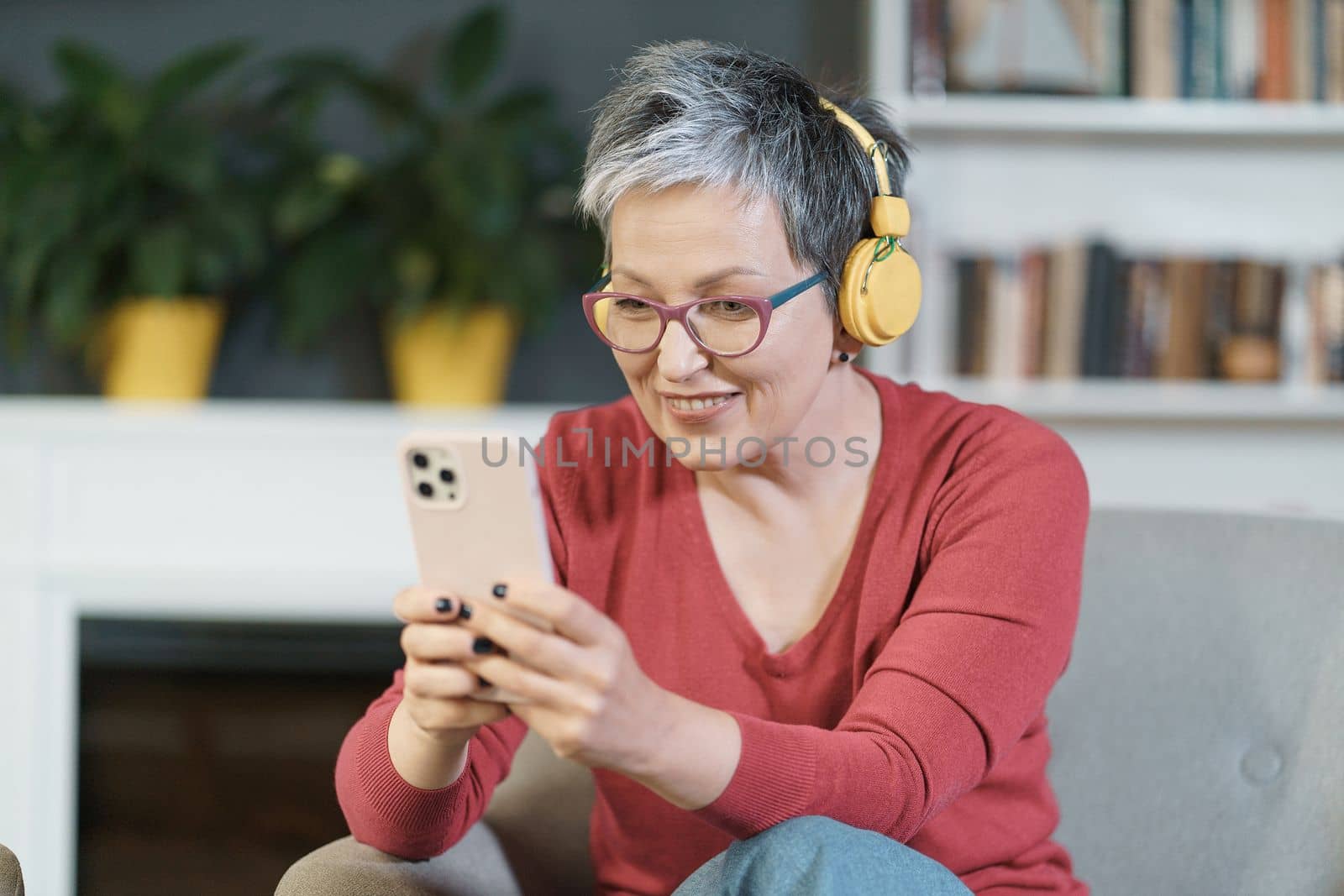 Mature woman wearing pink glasses and yellow headphones smiles while texting on her phone at home. The image depicts technology, communication, and leisure, while the woman's happy expression reflects her enjoyment and satisfaction with digital messaging.