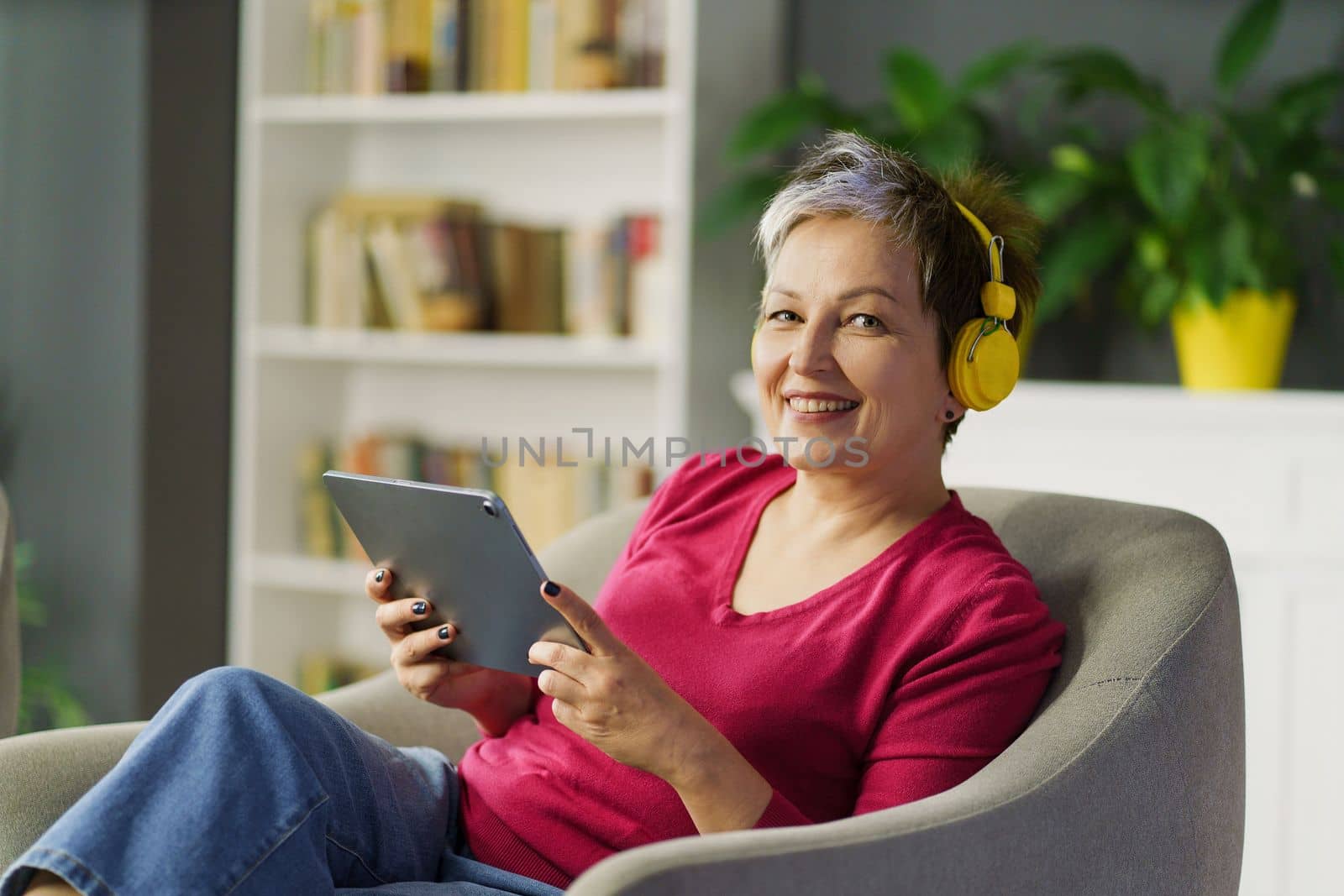 Mature woman smiles on a sofa, holding tablet PC and listening to music on headphones. She enjoys leisure, technology and music, feeling tranquil and blissful. High quality photo