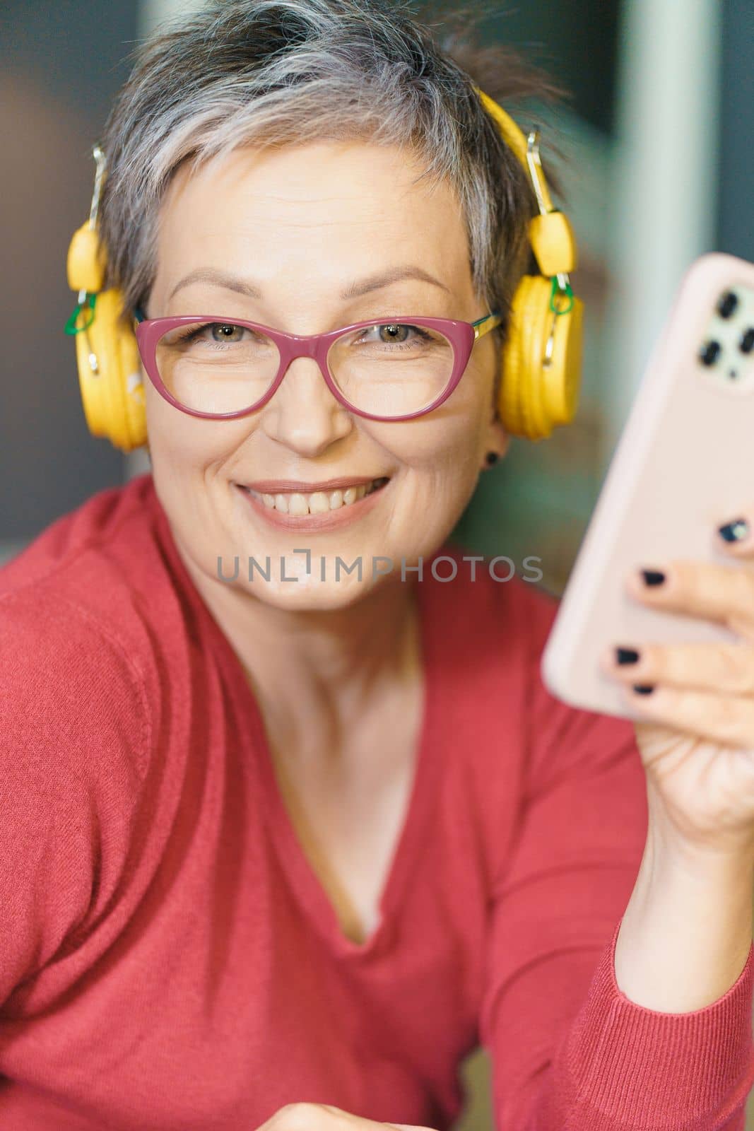 Mature woman wearing glasses, smiling and holding phone headphones on. Image portrays technology, communication, and entertainment, while woman's happy expression reflects her enjoyment and satisfaction with modernity. High quality photo