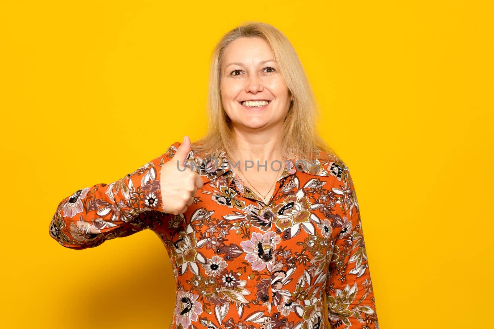 Photo portrait of nice caucasian woman with bright smile showing thumb up in patterned dress isolated on yellow colored background