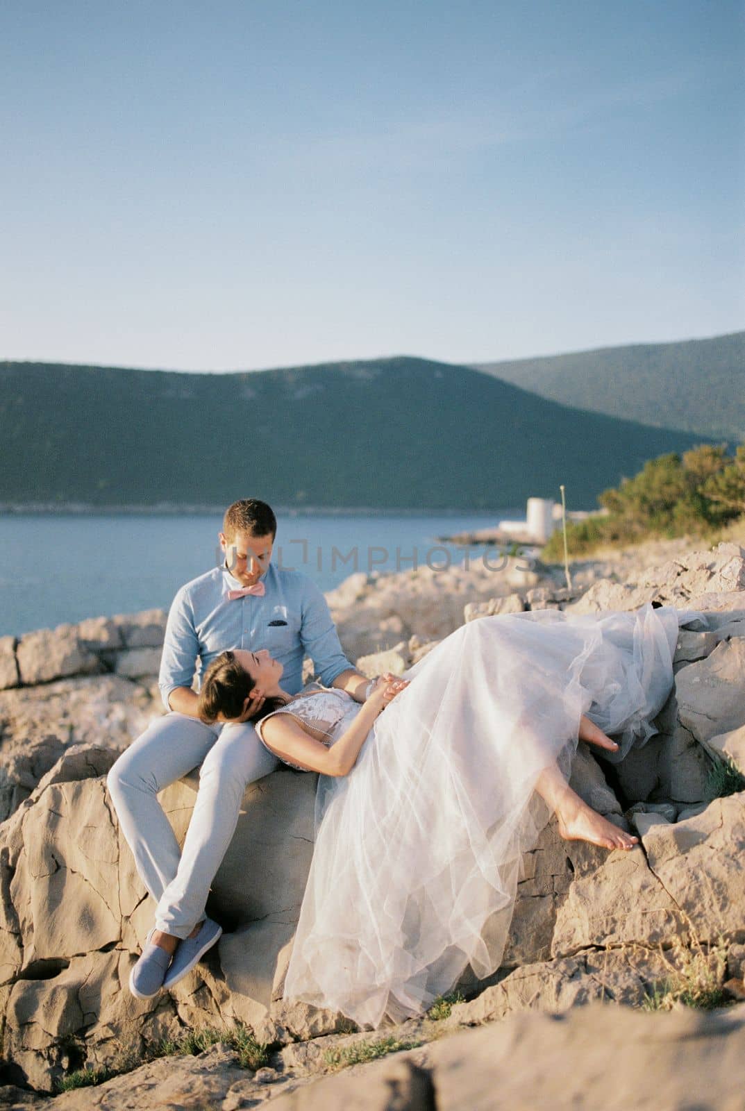Bride lies on the knees of the seated groom on a rocky seashore overlooking the mountains. High quality photo