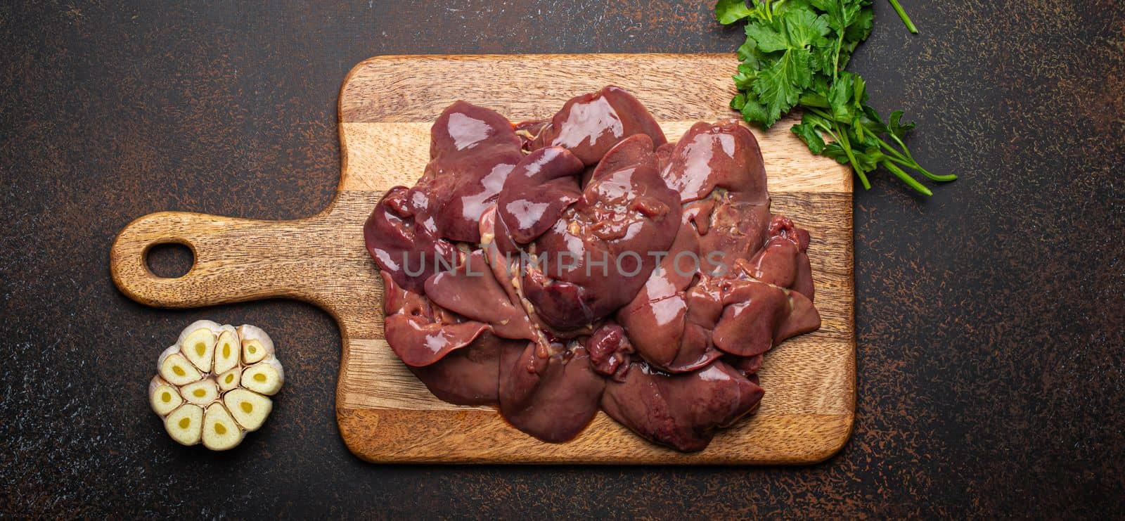 Raw chicken liver on wooden cutting board top view on dark rustic concrete background kitchen table with parsley and garlic. Healthy food ingredient, source of iron, folate, vitamins and minerals