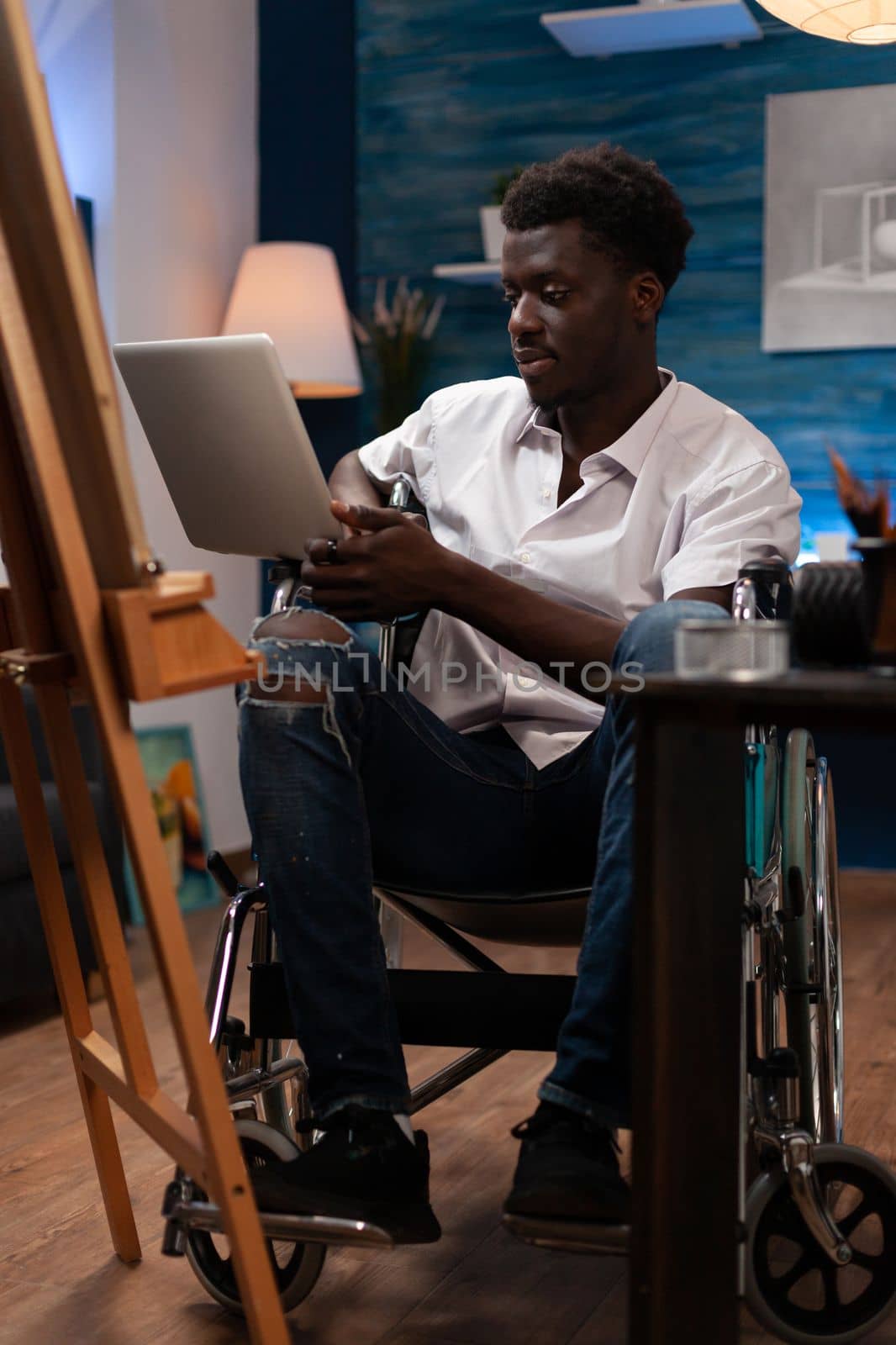 Person with artistic skills and disabilities taking online drawing courses with laptop. African american wheelchair user looking for inspiration on art web sites to create sketch masterpiece at atelier.