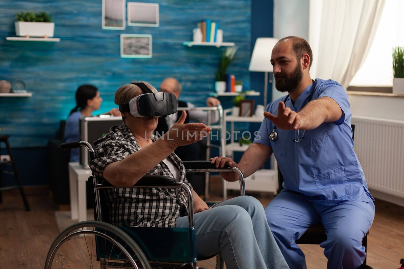 Elderly woman in wheelchair using virtual reality glasses in common room of nursing home, male nurse providing assistance and teaching how to use modern technology. Time for fun in care center.